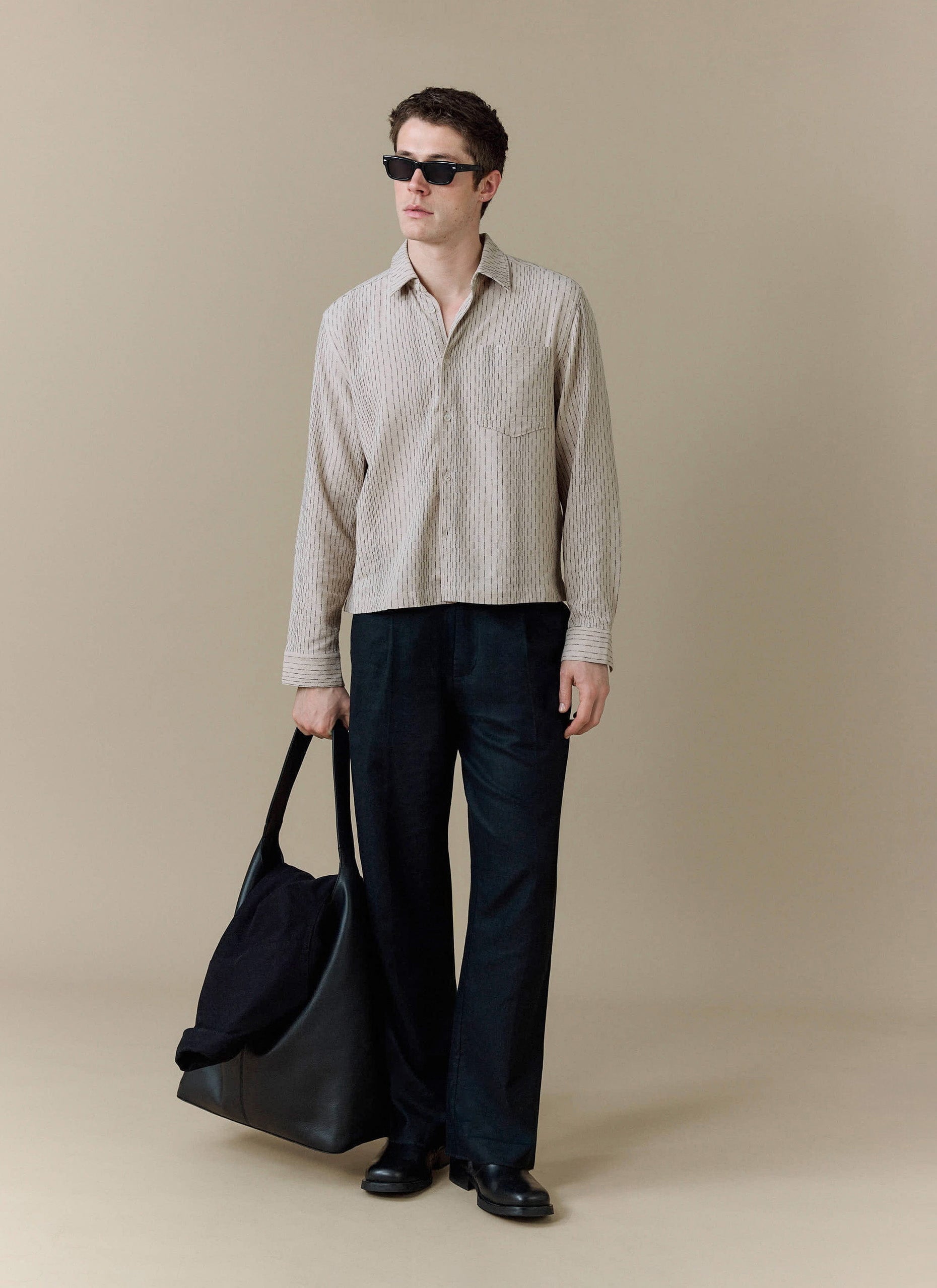 A model styles the Percival Leland Cuban Boxy Shirt in ecru with wide-leg black trousers, sunglasses and a large tote bag.