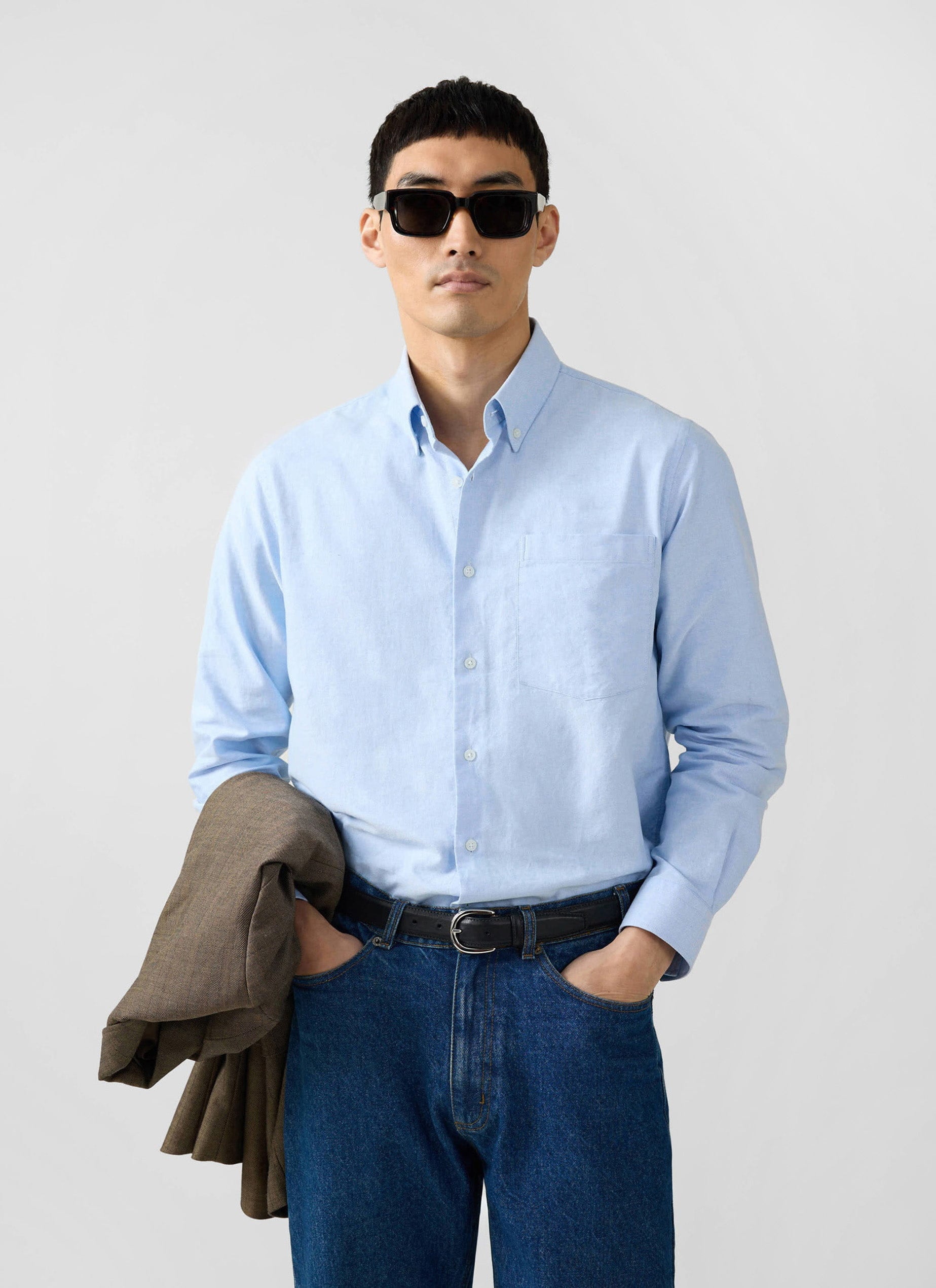 A model wears the Percival Oxford Regular Shirt in light blue, styled with dark jeans, sunglasses and a brown jacket.