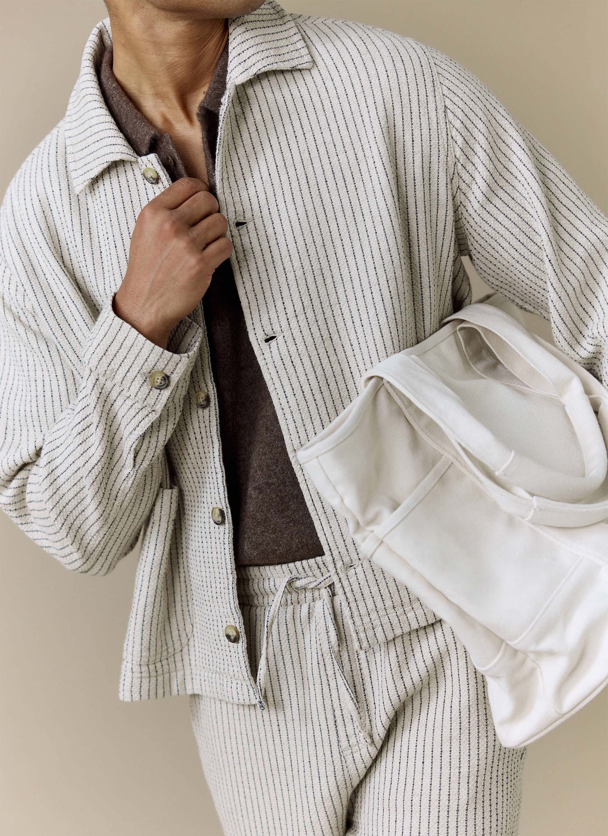 A close-up of a model wearing the Percival Sorrento Boxy Overshirt in ecru, layered over a brown top and carrying a tote bag.