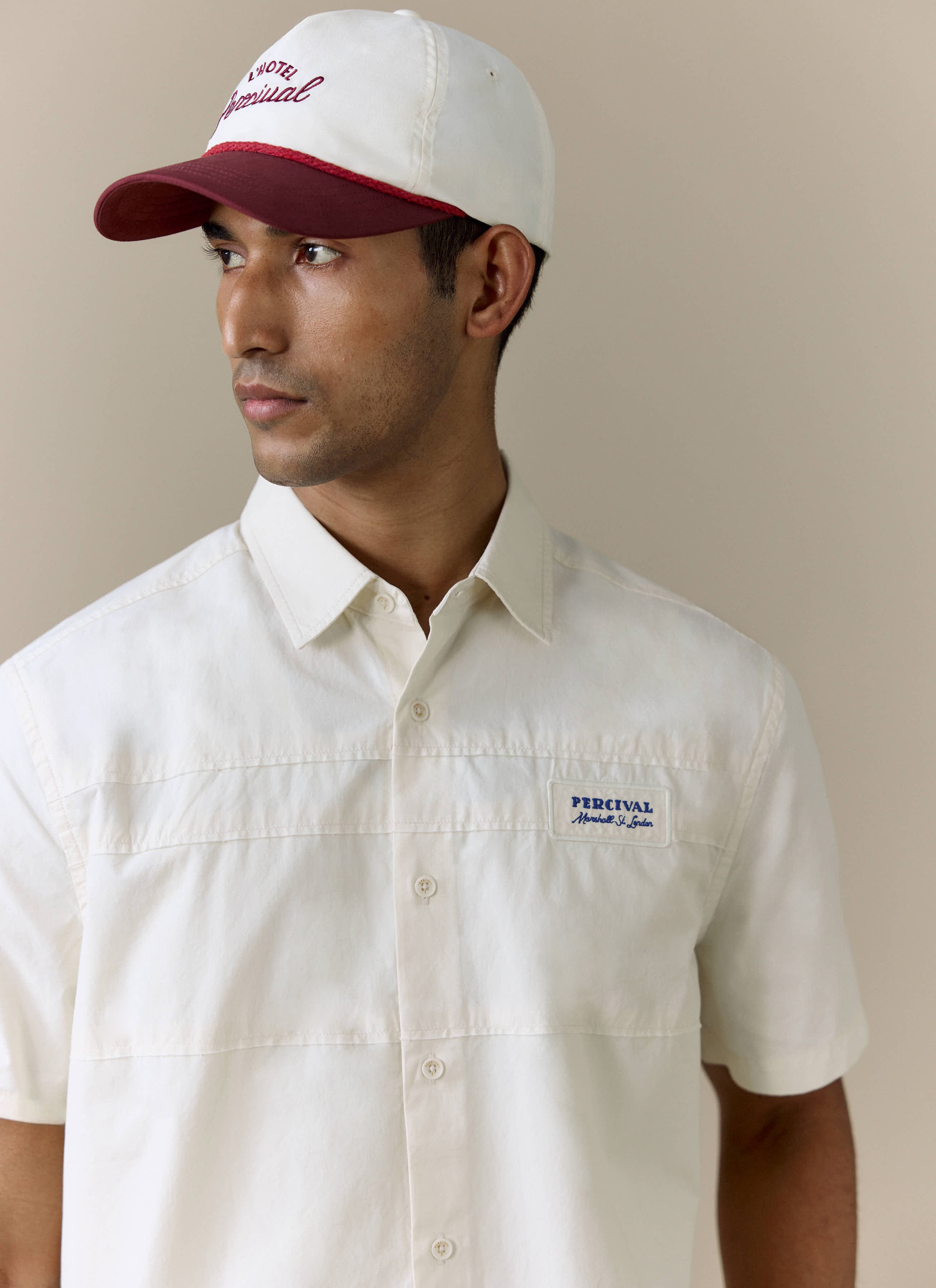 A model wears the Percival Harlow Boxy Shirt in ecru cotton, showing the blue embroidered chest badge.