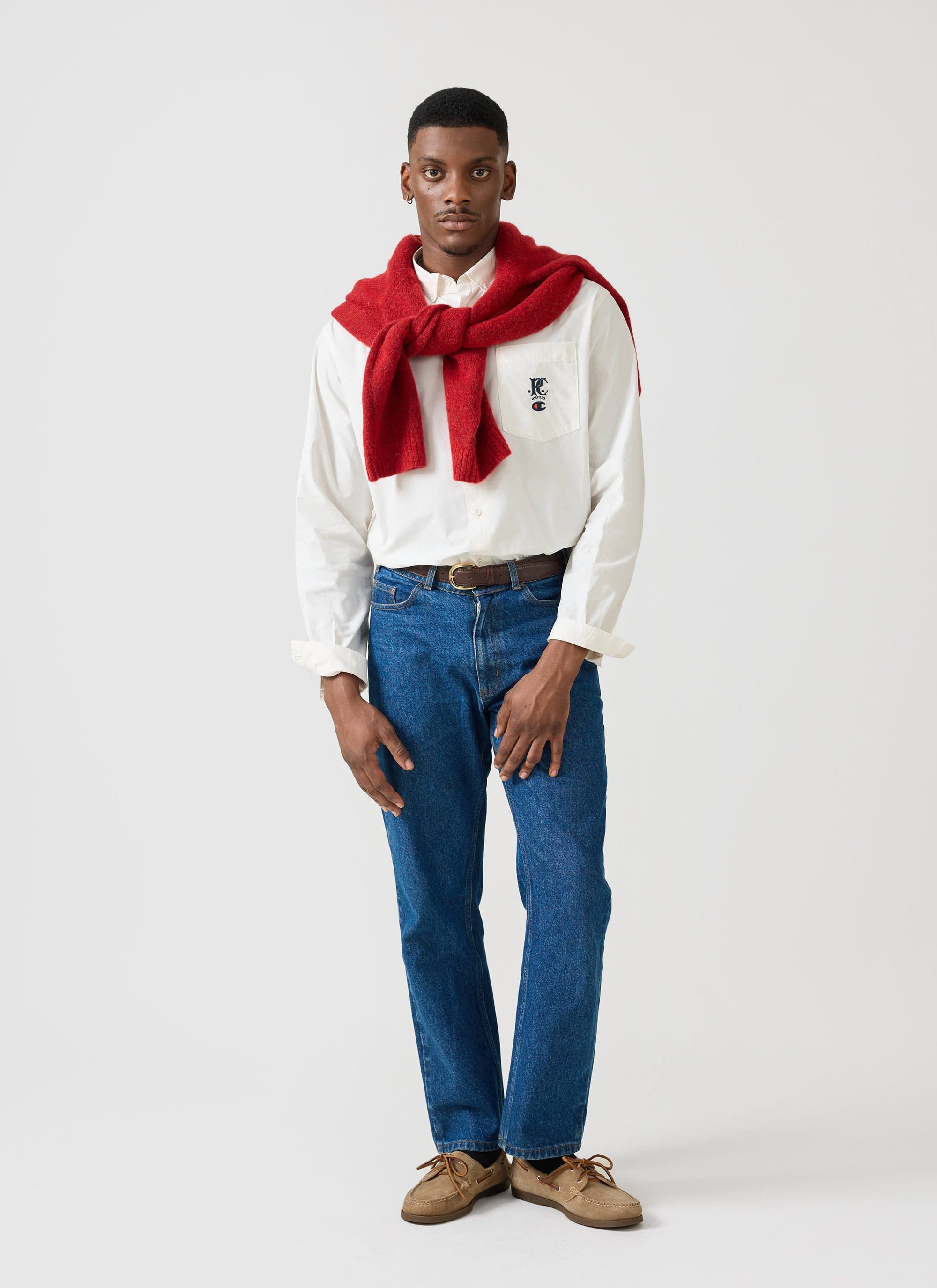 A model wears the ecru Champion x Percival Oxford shirt, styled with blue jeans and a red jumper over his shoulders.