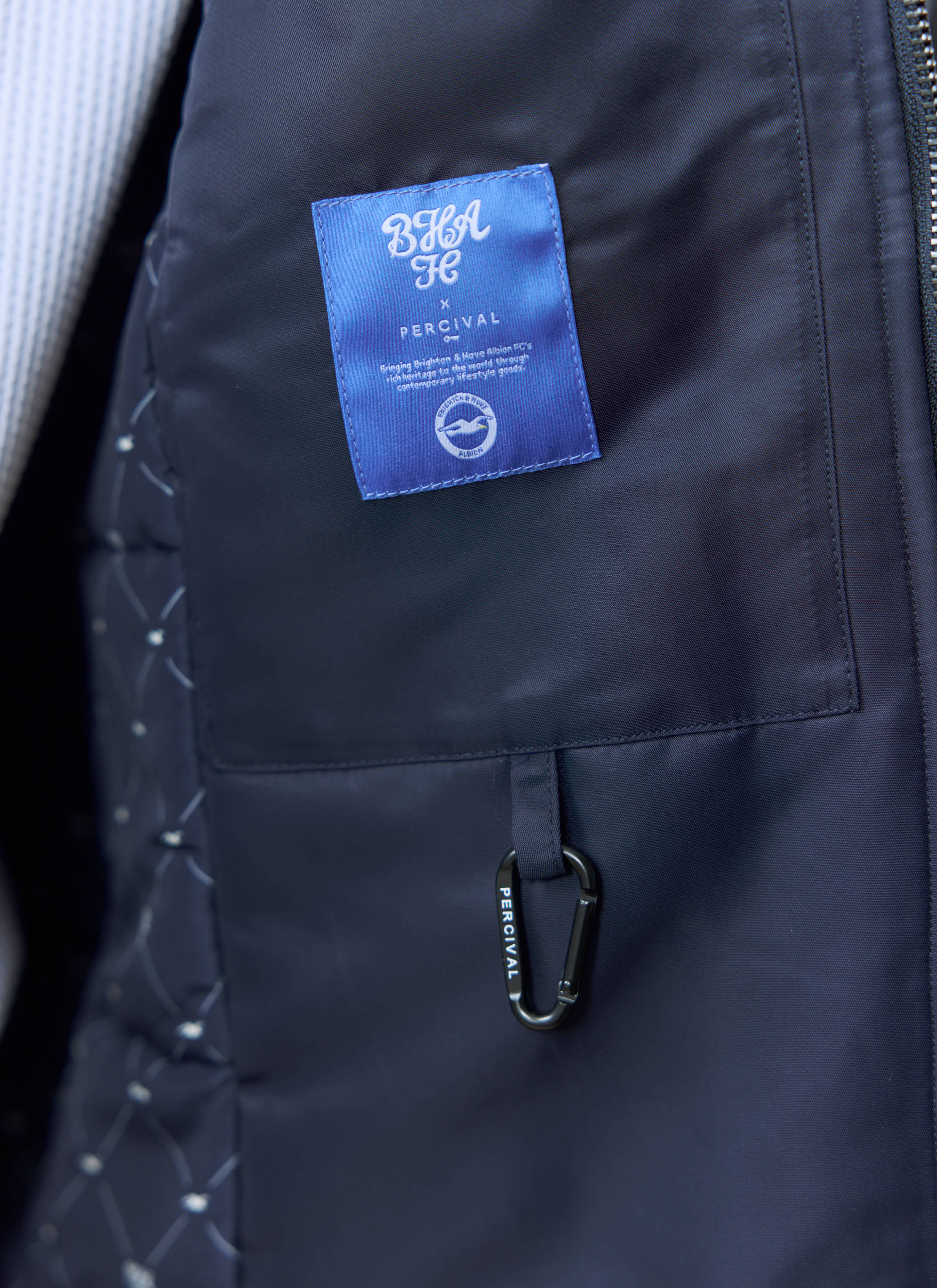A close-up of the internal pocket and blue collaboration label of the navy Percival x Brighton & Hove Albion Coach Jacket.