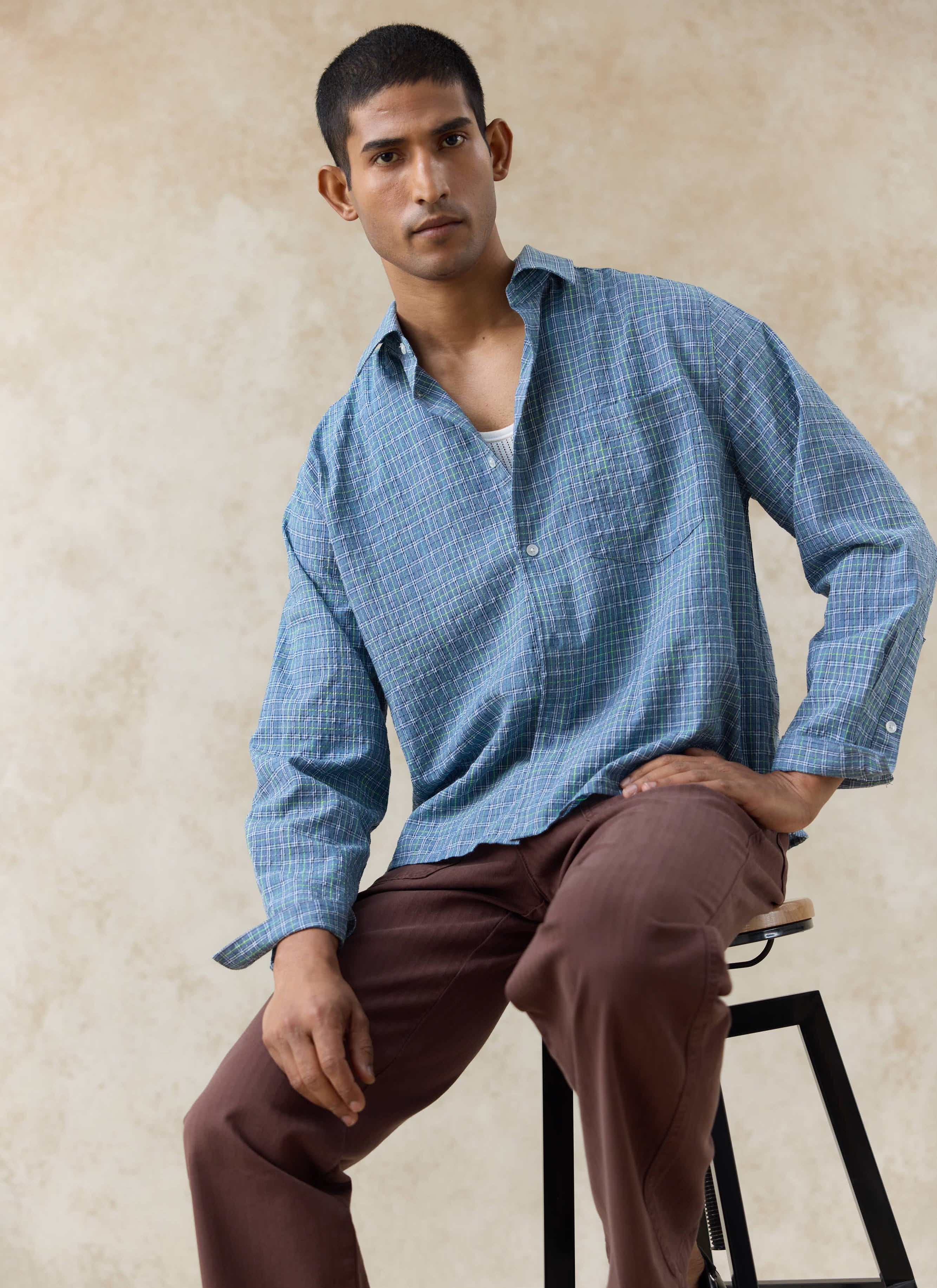 A model wearing the Percival Hinsley Checked Boxy Shirt in blue cotton, paired with brown trousers and a white vest.
