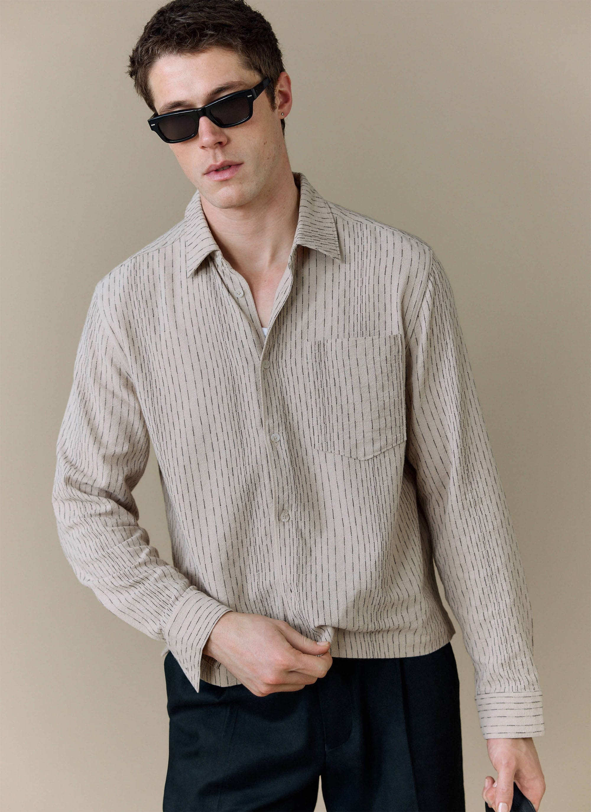 A model wears the ecru cotton Leland Cuban Boxy Shirt from Percival, styled with black trousers and sunglasses.