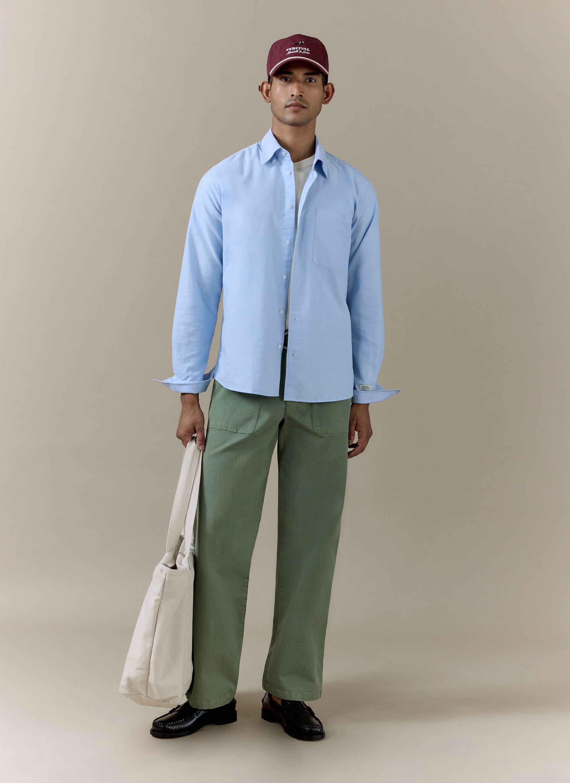 Model wearing the sage green cotton Lungo Herringbone Straight Trousers by Percival with a blue shirt and tote bag.