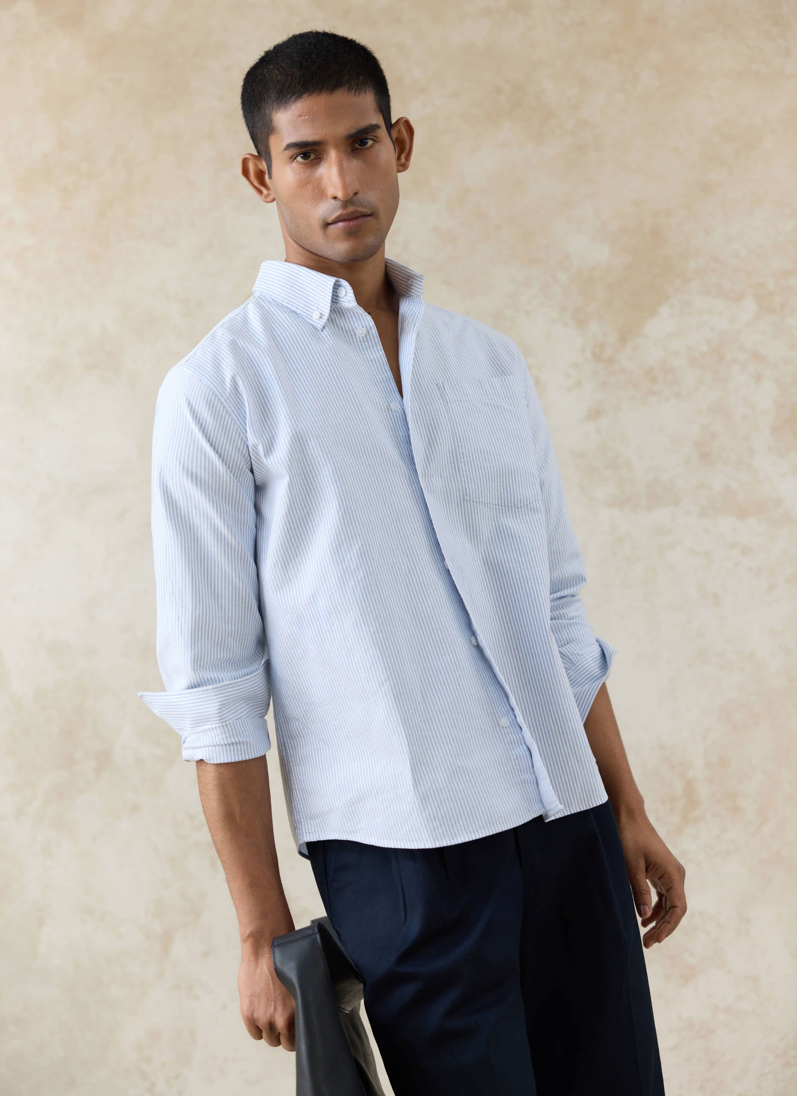Model wears the Percival blue pinstripe Oxford shirt in cotton with rolled sleeves and navy trousers.
