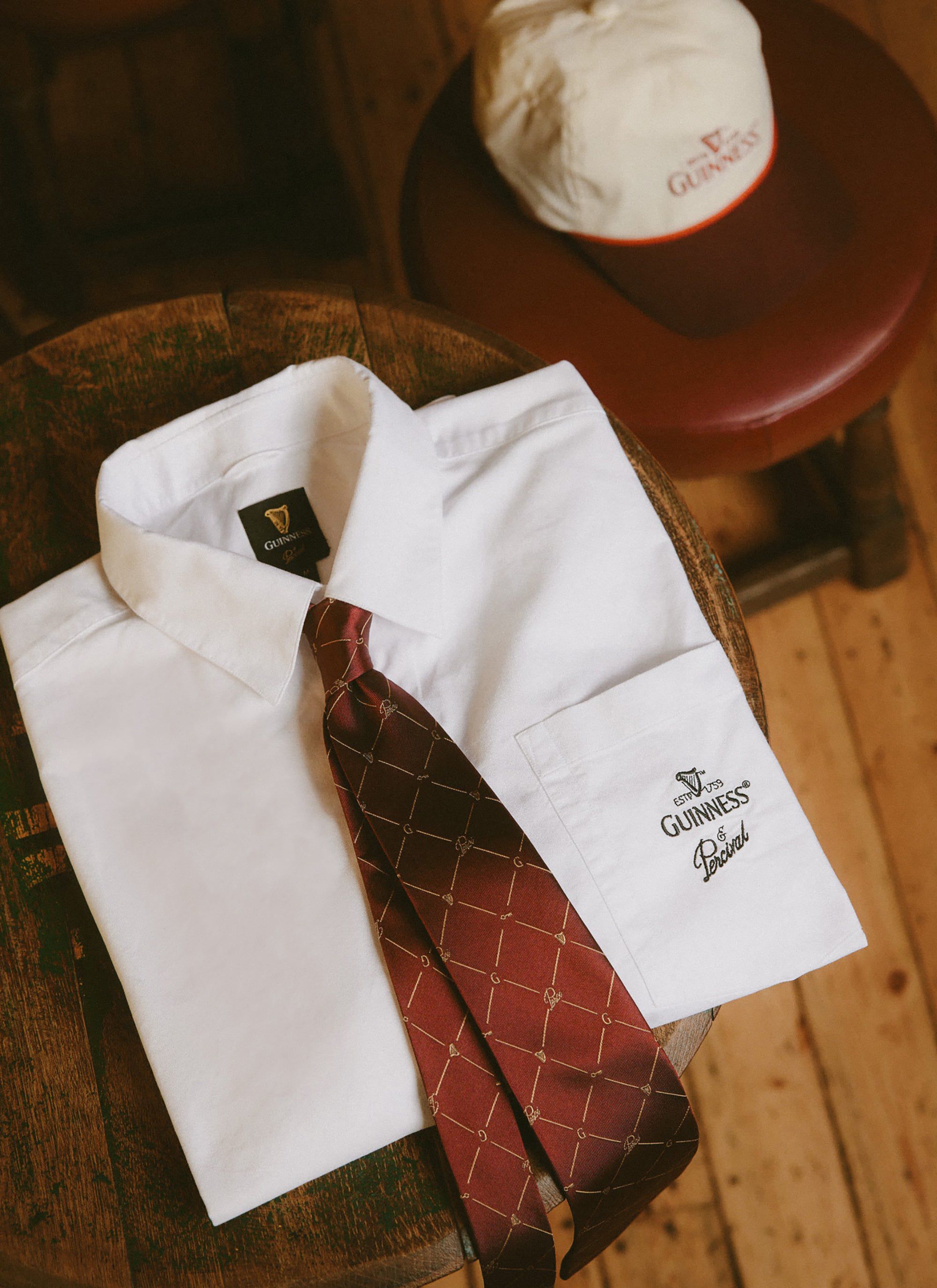 The Percival x Guinness Button Down Boxy Shirt in white, laid on a wooden stool with a patterned tie and branded cap.