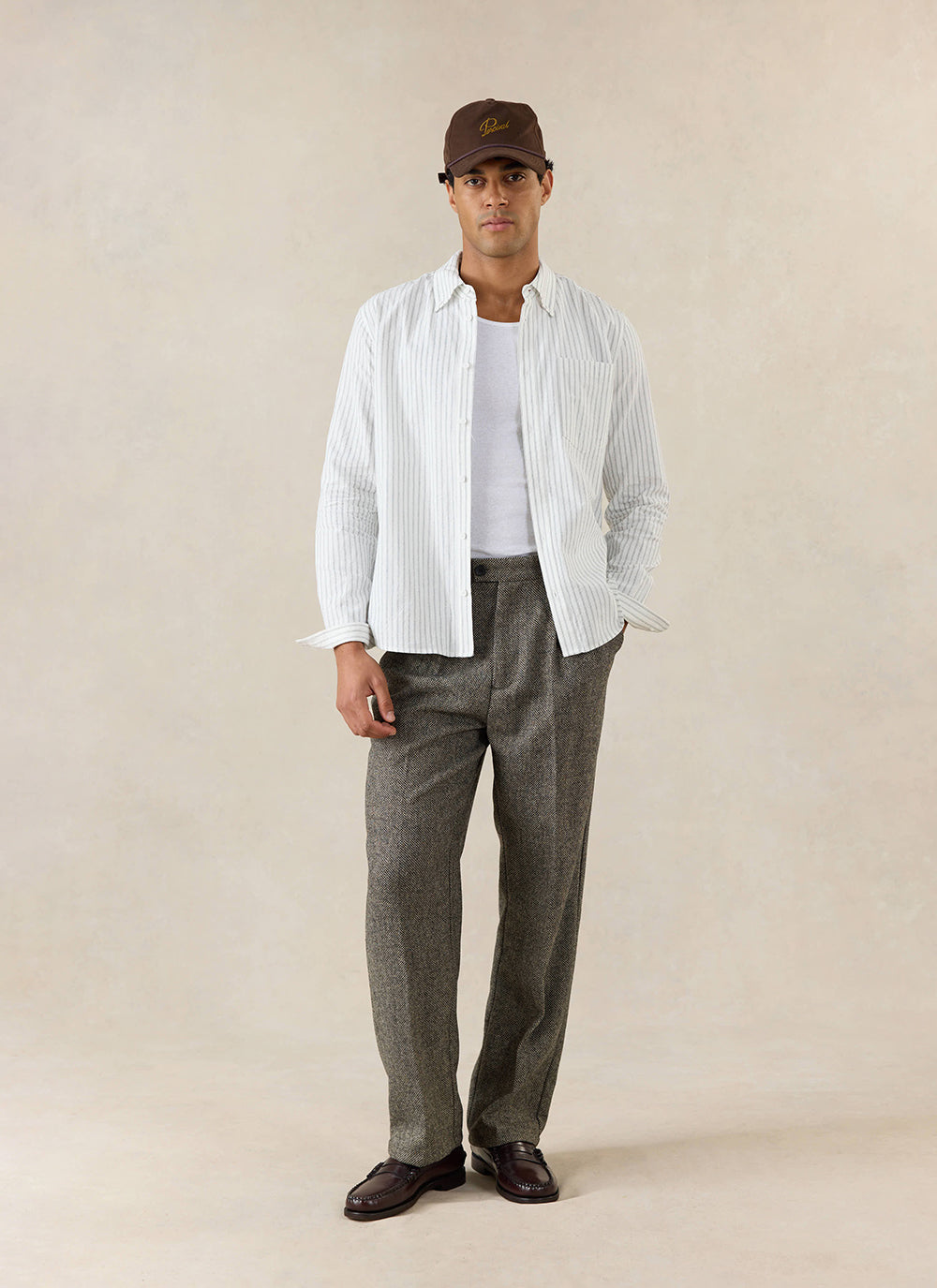 A model wears the Percival Blake Double Pleated Tailored Trousers in grey birdseye wool, a striped shirt and brown cap.