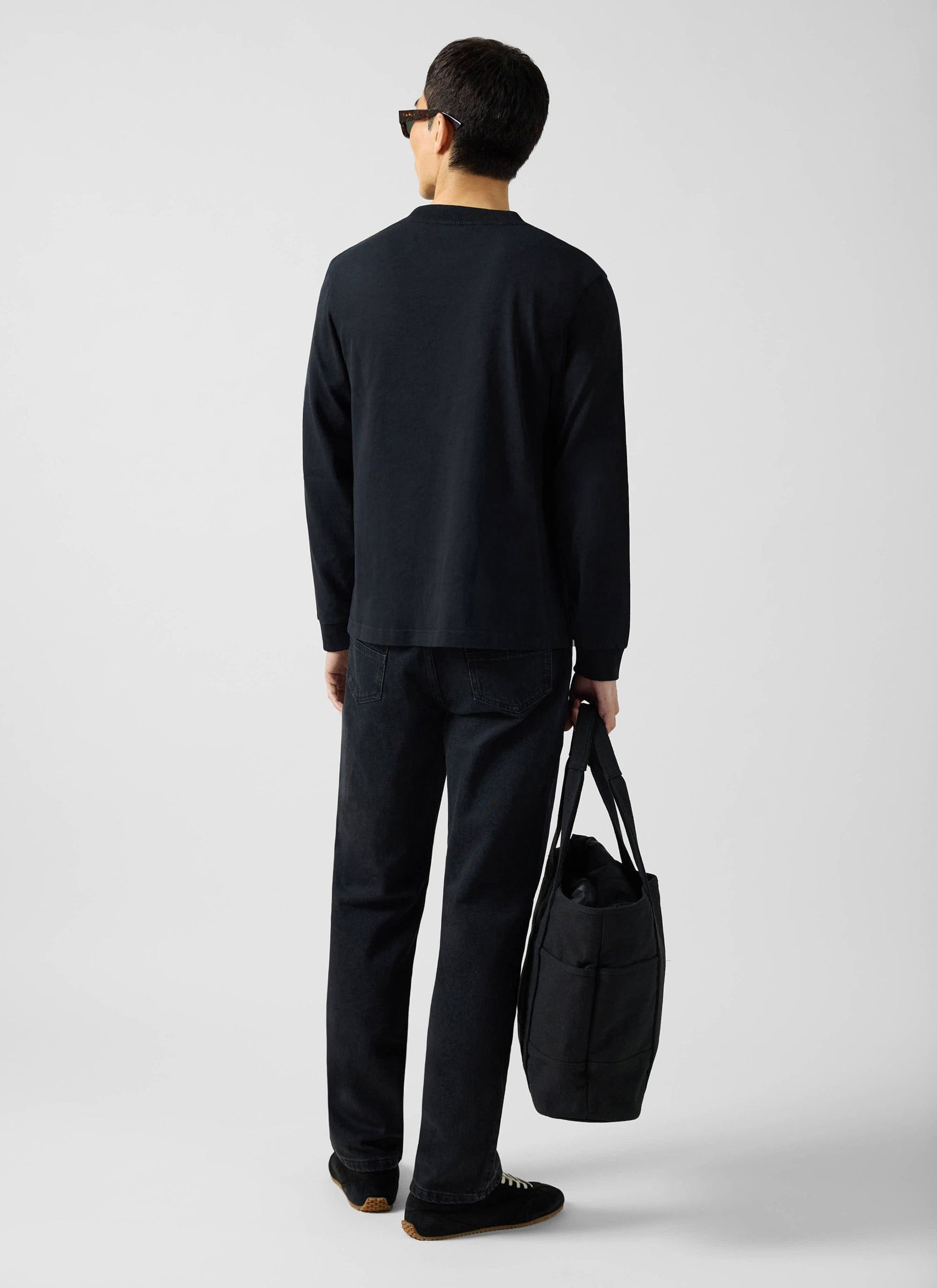 Back view of a model wearing the Percival black long sleeve organic cotton t-shirt, styled with black jeans and a tote bag.
