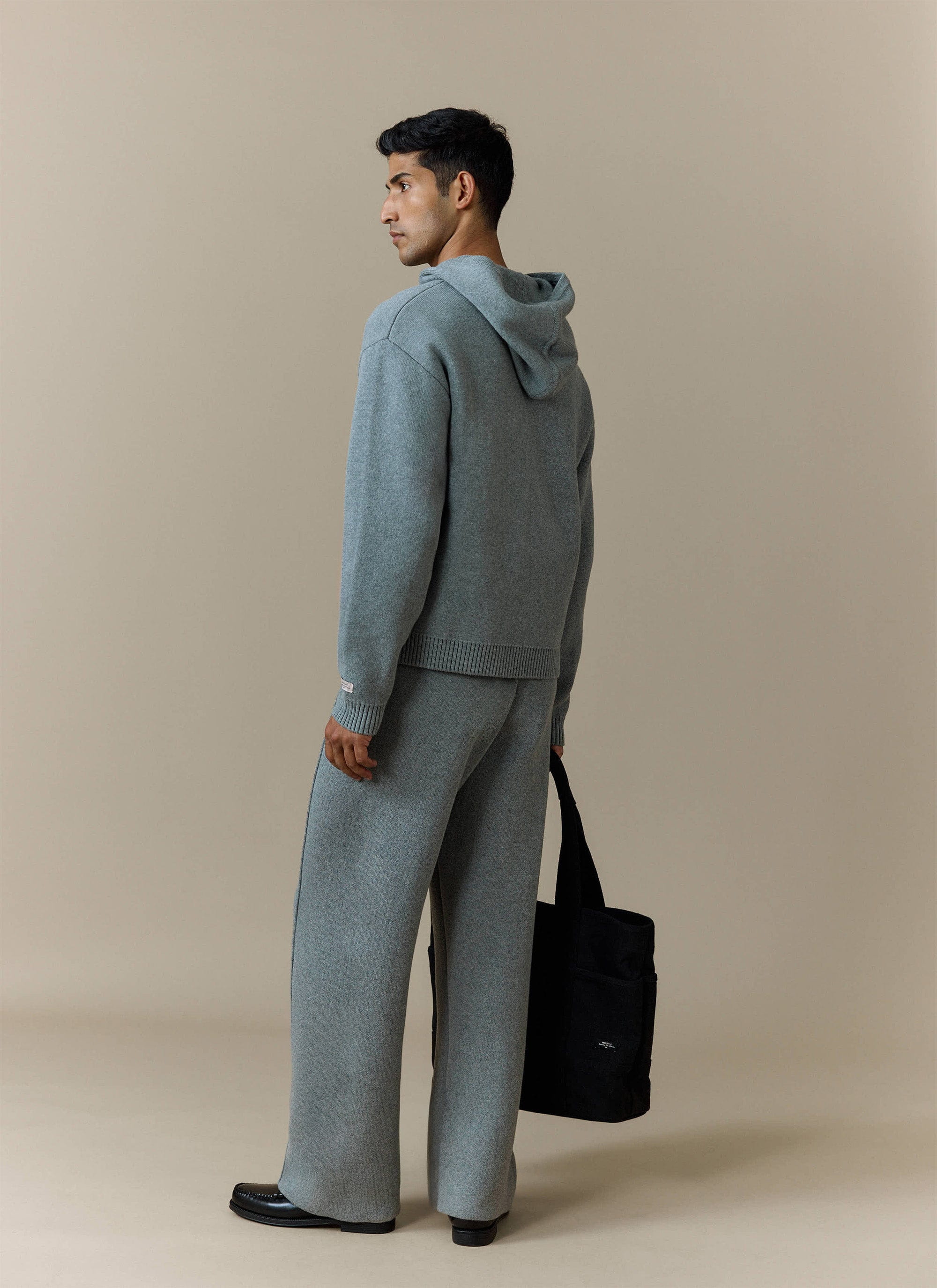 A model wears the grey Chronicle Knitted Hoodie from Percival, styled with matching wide-leg trousers and a black tote bag.