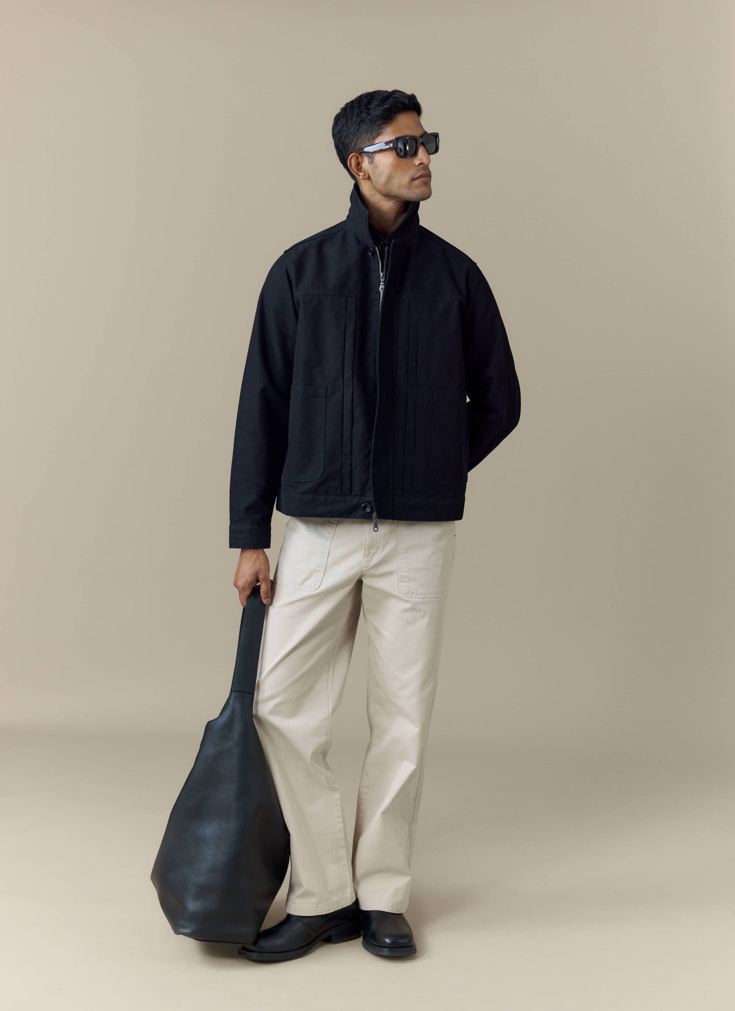 A model wears the black cotton Ernst Jacket from Percival, styled with cream trousers, sunglasses and a black bag.