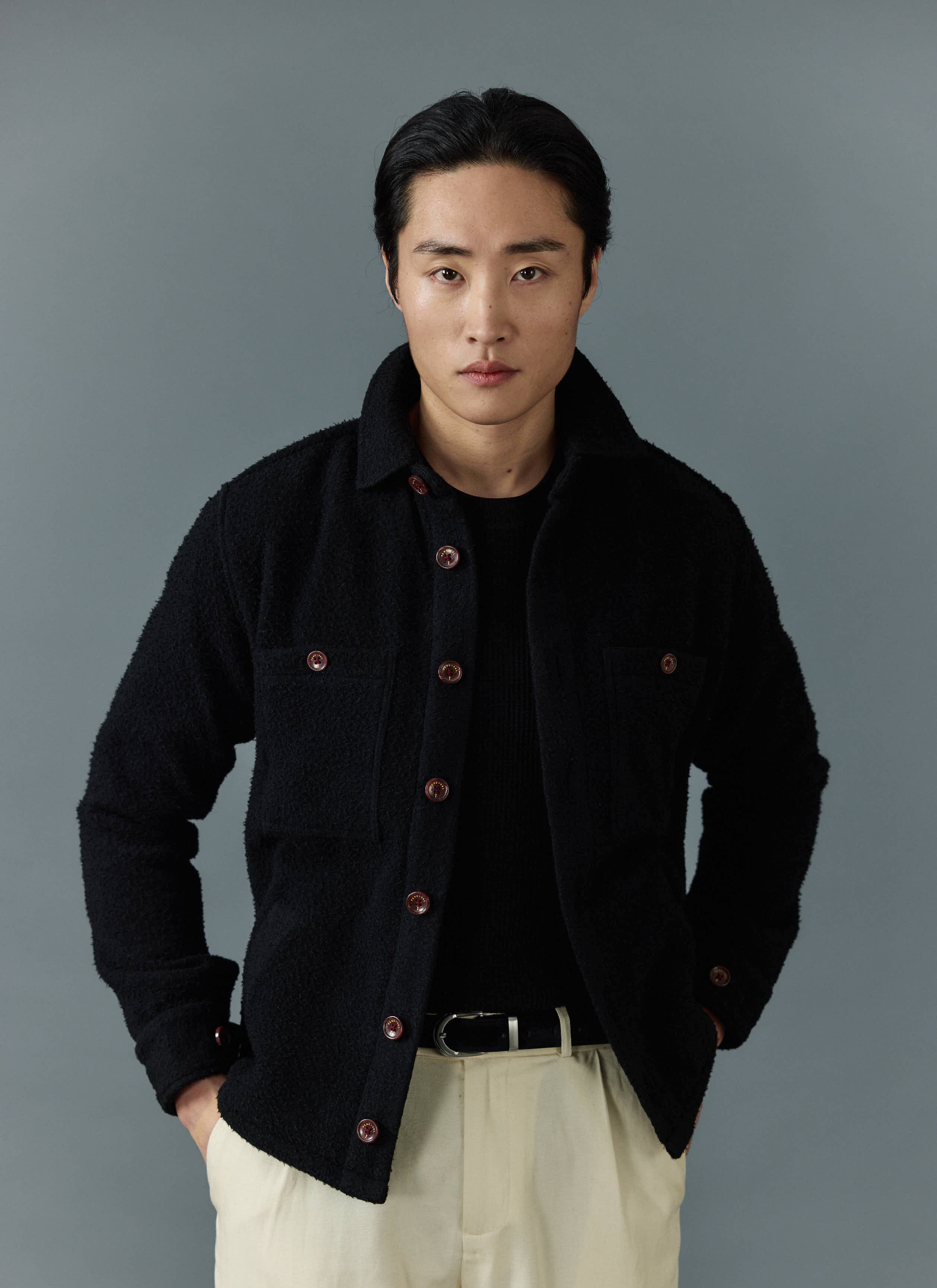 A model wears the Percival black Casentino wool Blanket Overshirt with cream trousers against a grey background.