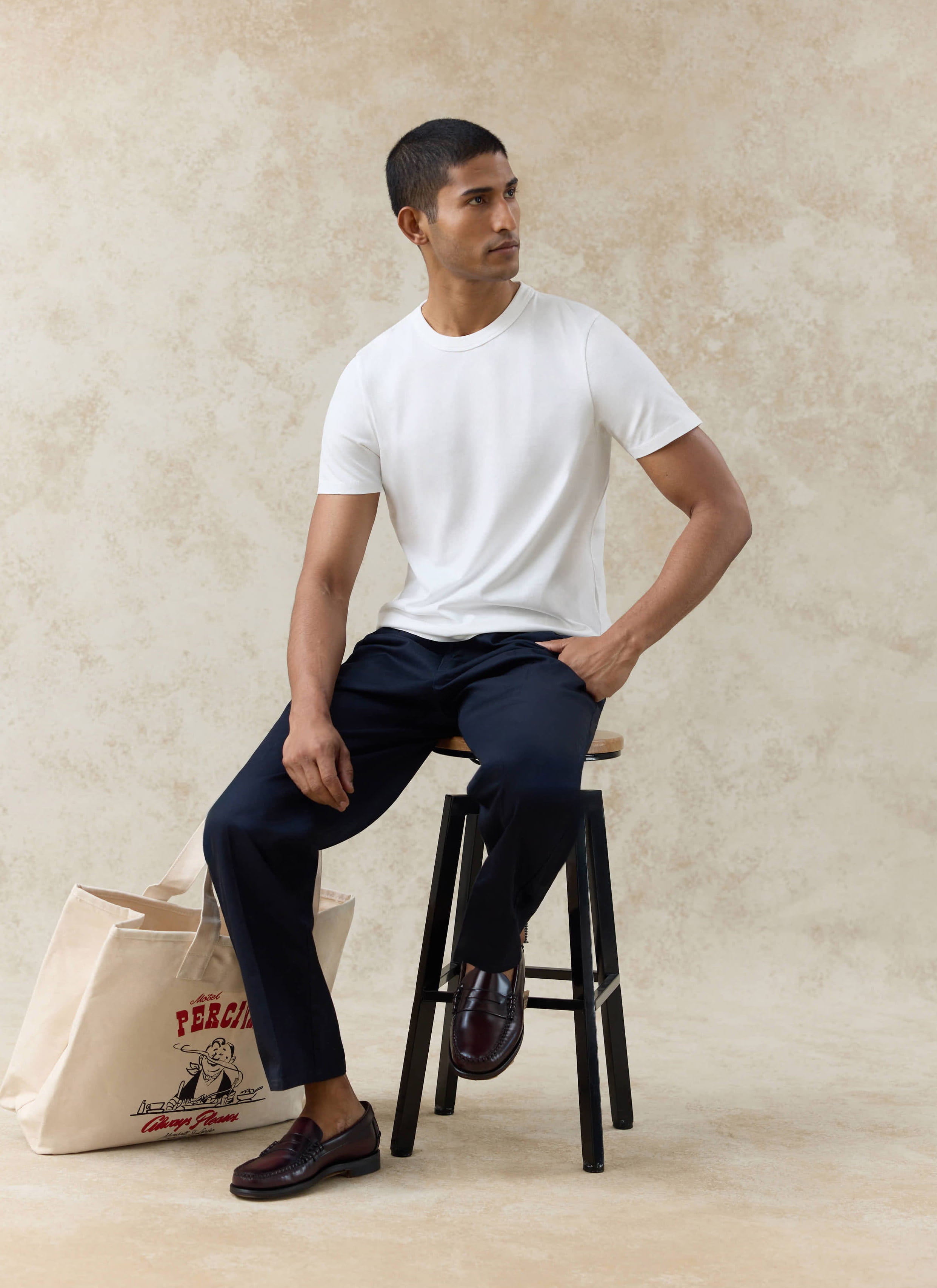 Model in navy Percival Blake double pleated cotton trousers and an off white Austin slub T-shirt, sitting on a stool.