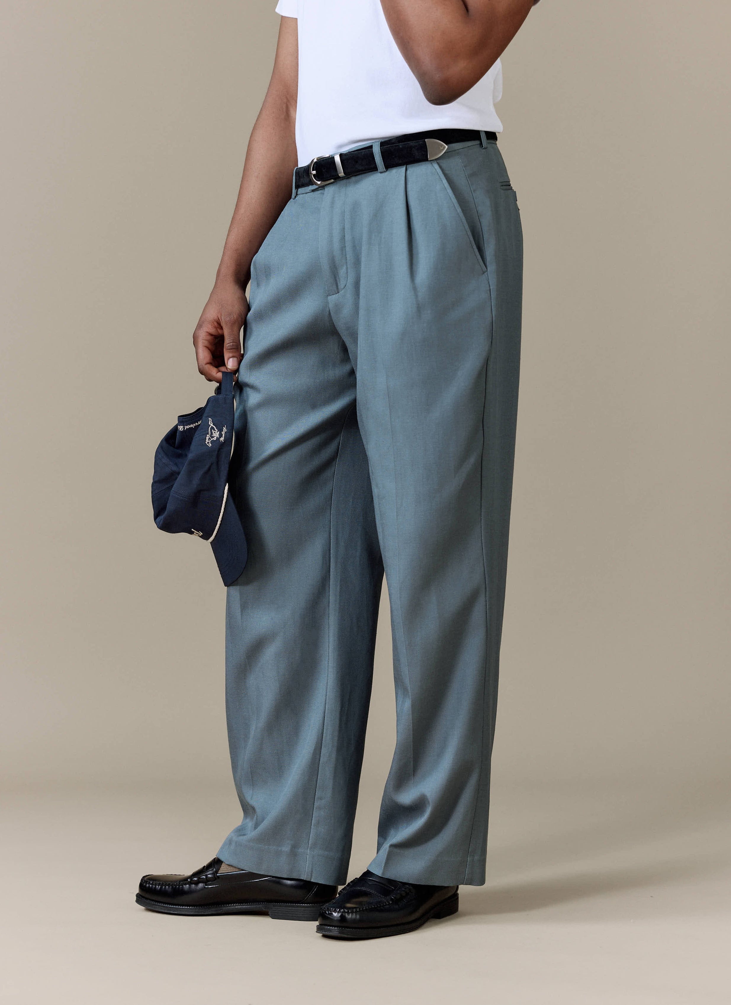 A model wears the Percival Pleated Leg Trousers in slate blue, styled with a white t-shirt and black loafers.