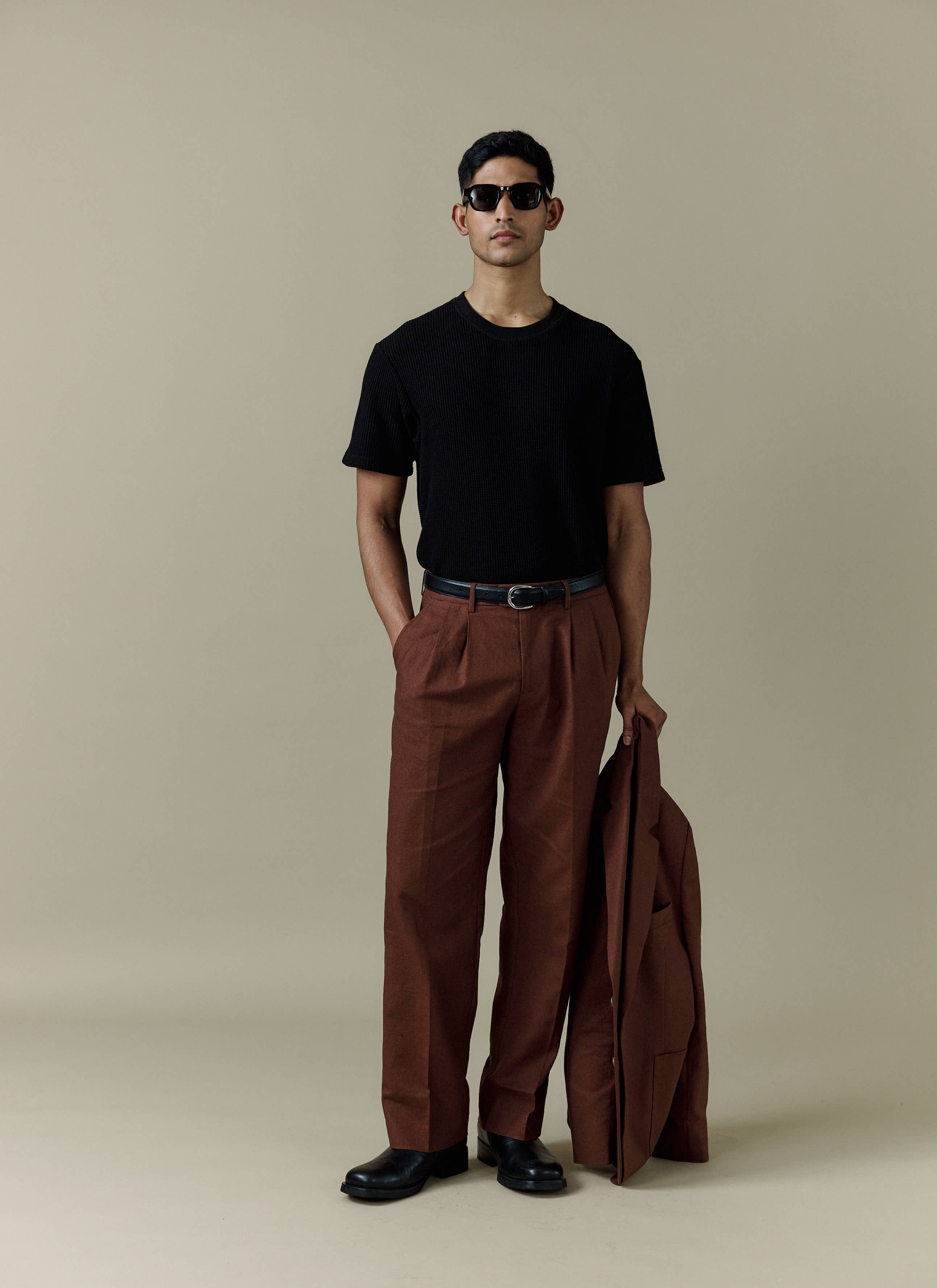 A man models the Percival brown linen Pleated Tailored Trousers, styled with a black t-shirt and matching suit jacket.