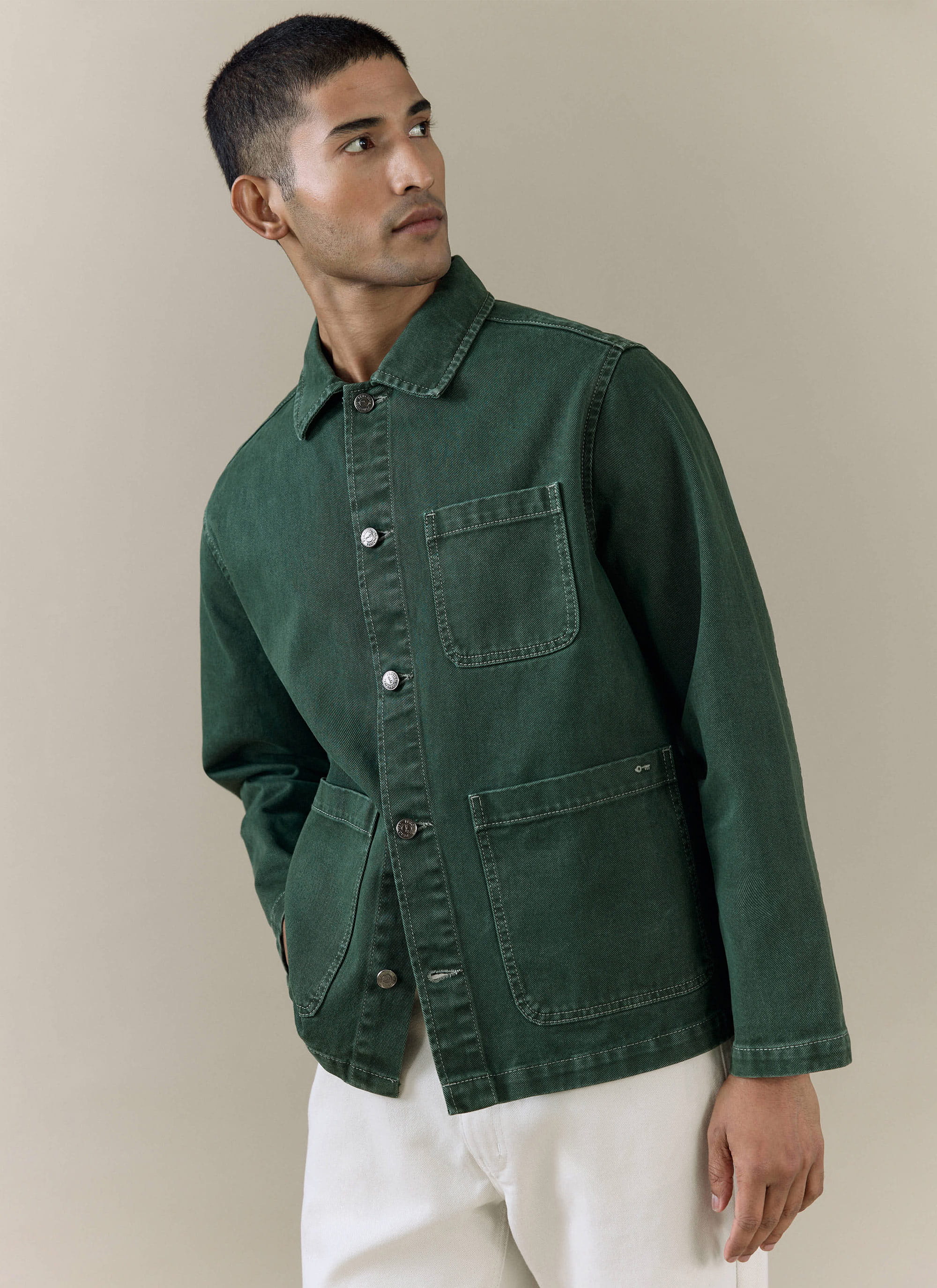 A model wears the forest green Percival Vintage Wash Oliver Chore Jacket in cotton canvas with white trousers.