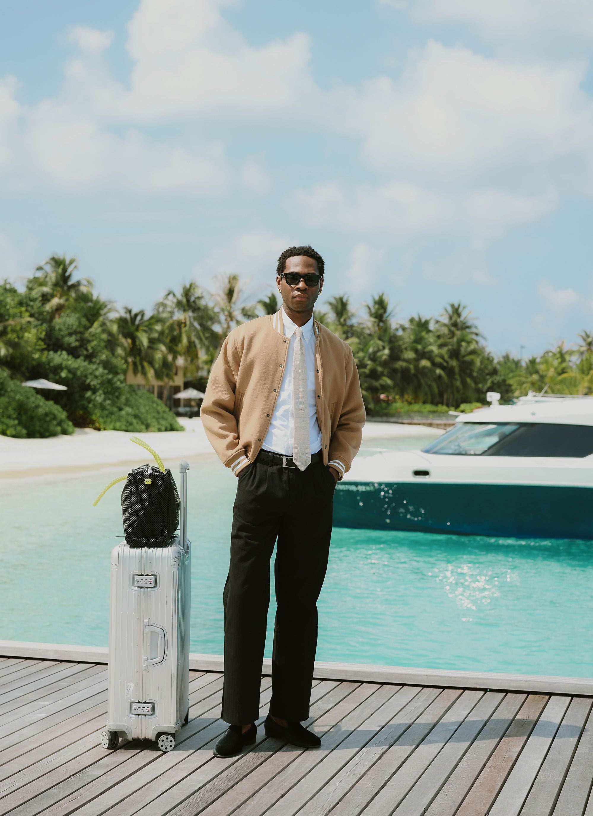 A man models the tan wool Percival Puglia Varsity Bomber Jacket on a pier next to a yacht and luggage.