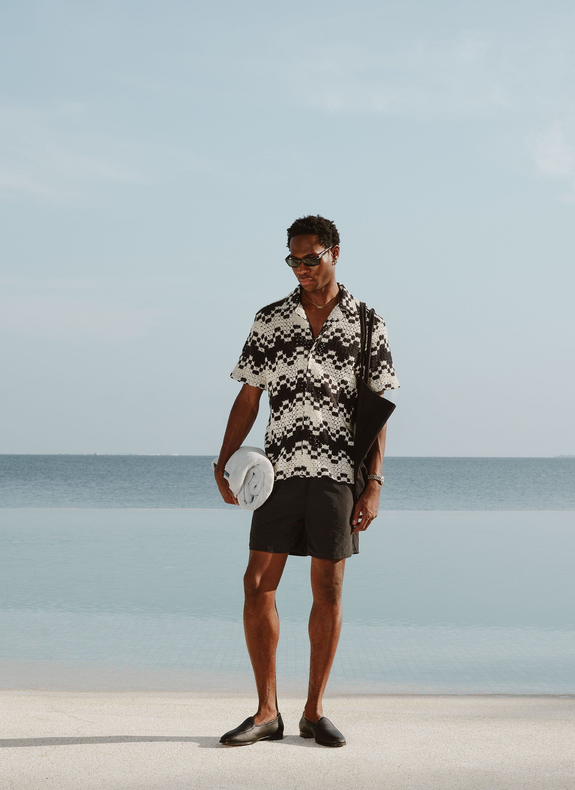 A man stands by the sea wearing the Percival Naples Crochet Cuban Shirt in ecru, paired with black shorts and loafers.