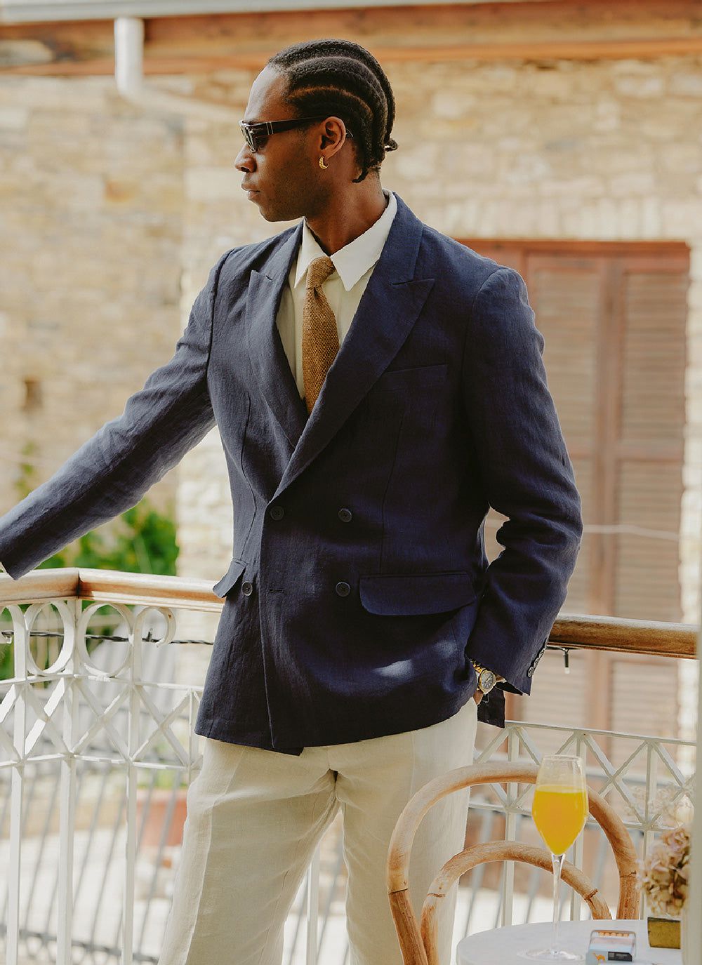 A man on a balcony wears the Percival navy linen tailored double breasted blazer with cream trousers and a brown tie.