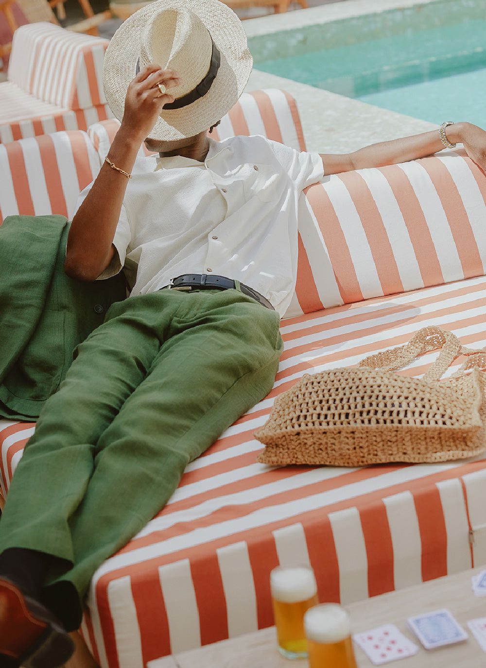 A model relaxes on a striped sun lounger wearing Percival's forest green tailored linen trousers with a white shirt and hat.