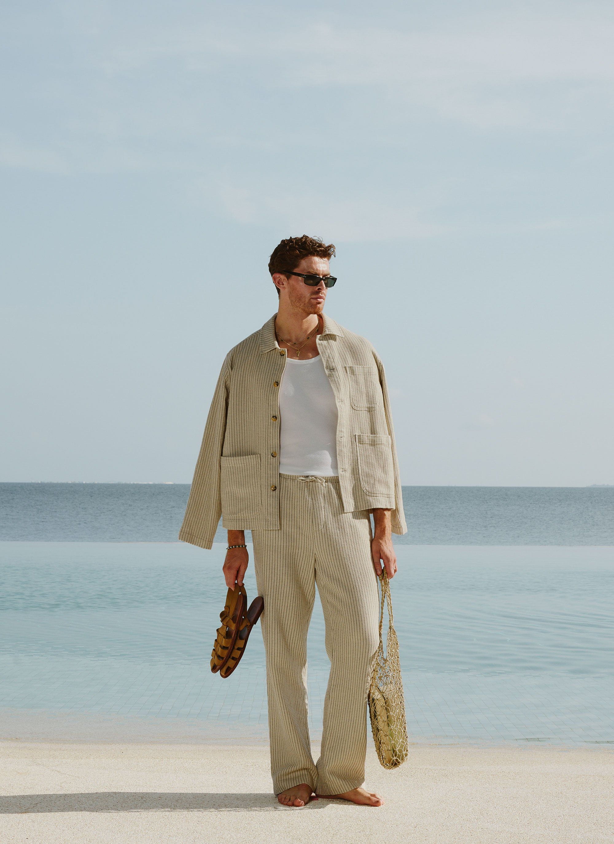 A model on a beach wears the Percival Sorrento Linen Trousers in ecru with the matching co-ord jacket.