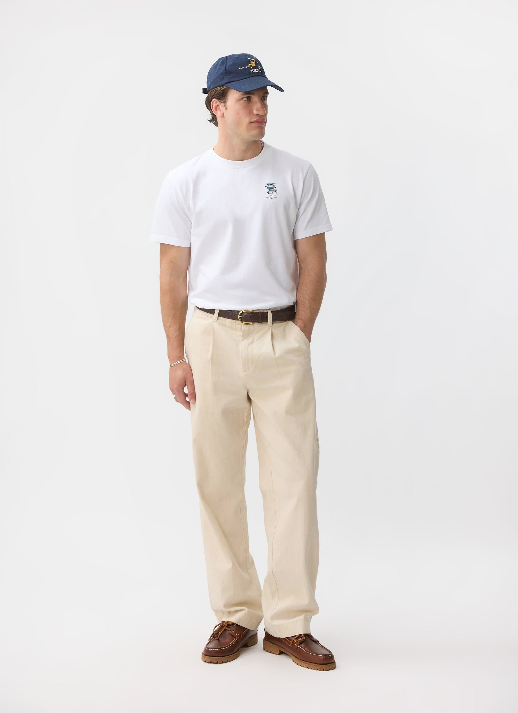 A man models the white Percival Duke T Shirt, styled with cream trousers, brown boat shoes, and a navy cap.