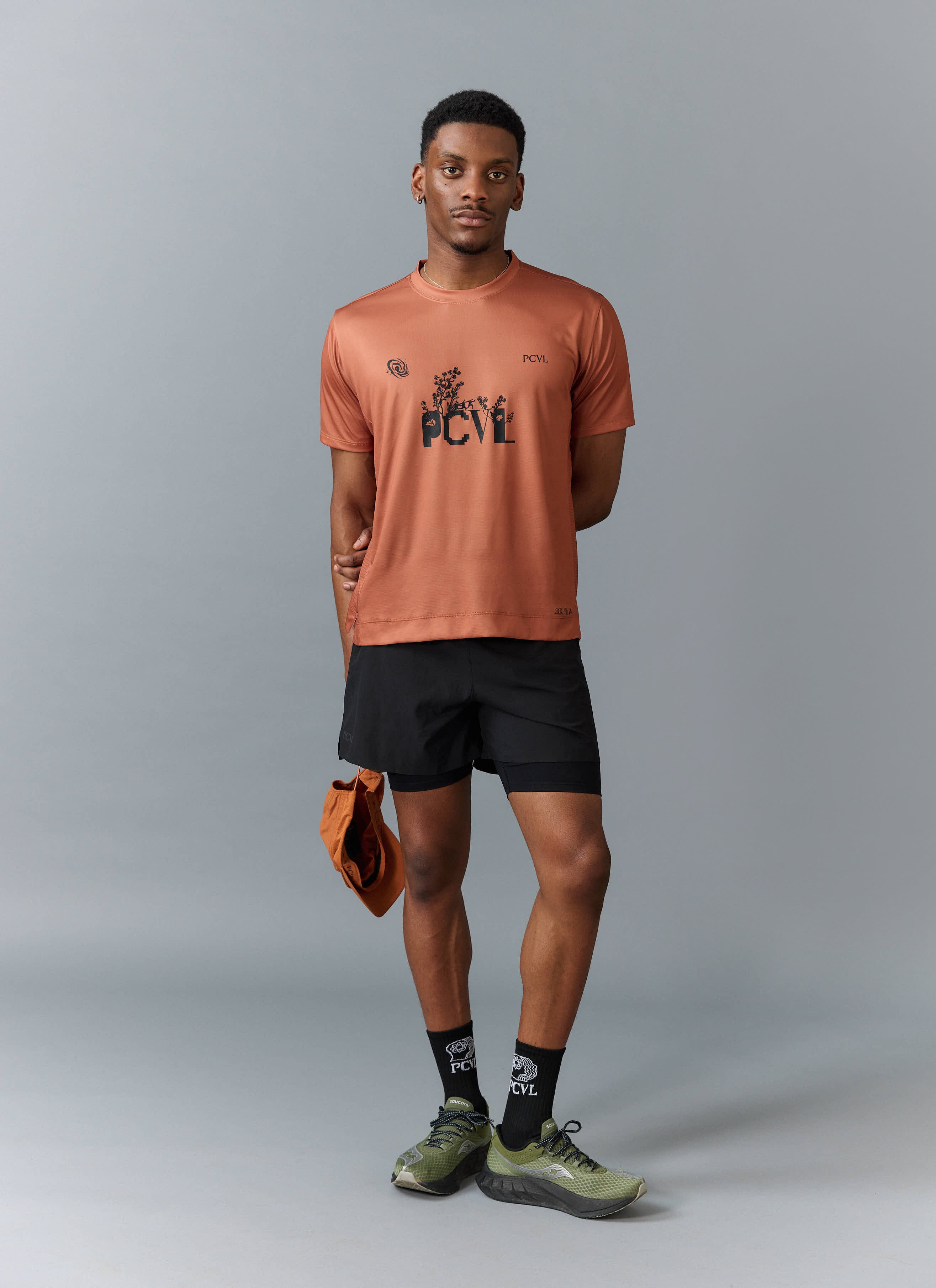 A model wears the rust PCVL Printed Performance Top from Percival, styled with black shorts, socks, and a matching cap.