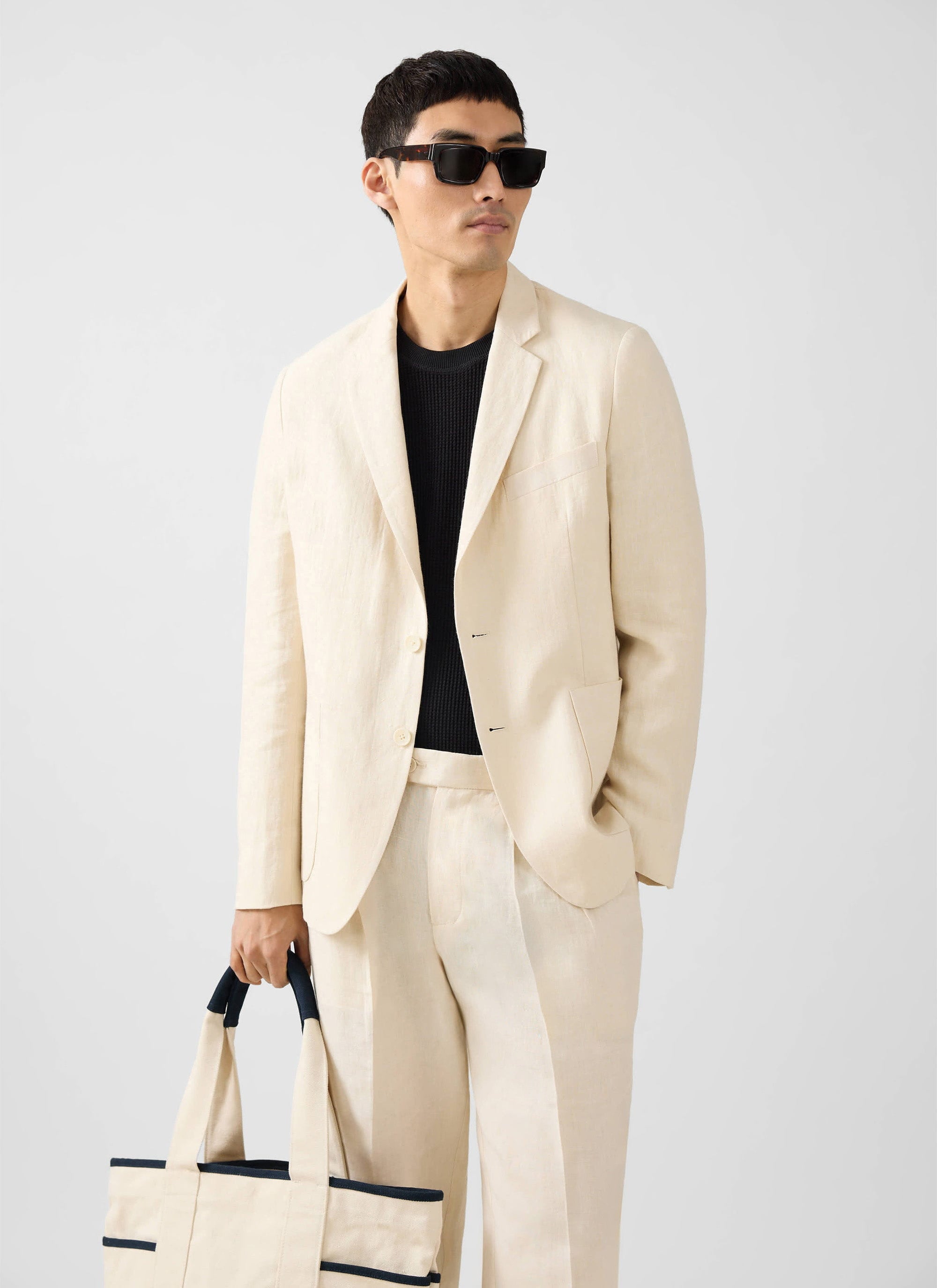 A model wears the Percival Tailored Linen Blazer in cream with matching trousers, a black jumper, sunglasses and a tote bag.