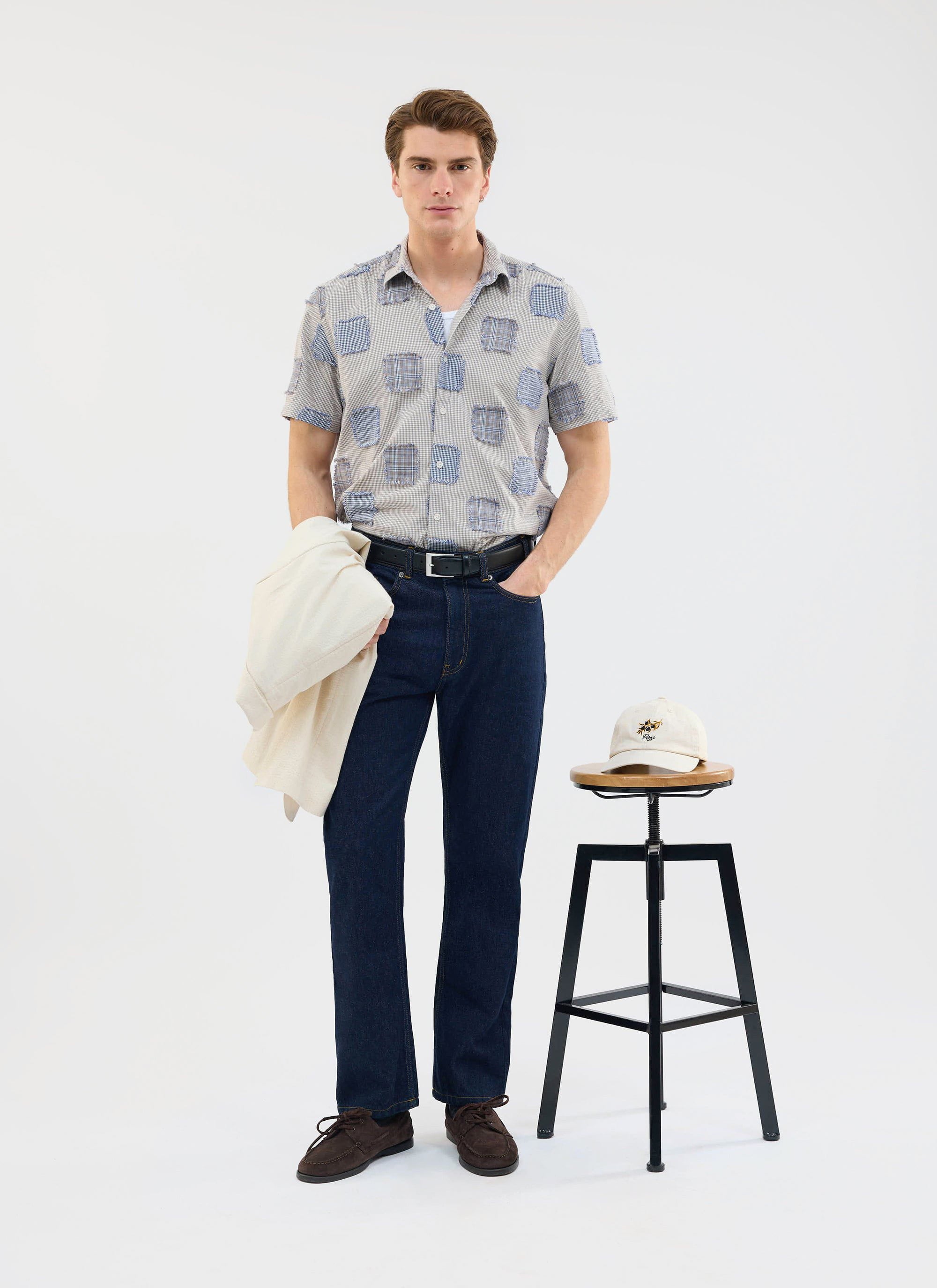 A model wears the Percival Bruno Boxy Clerk Shirt in grey, a patchwork design, paired with dark denim jeans.