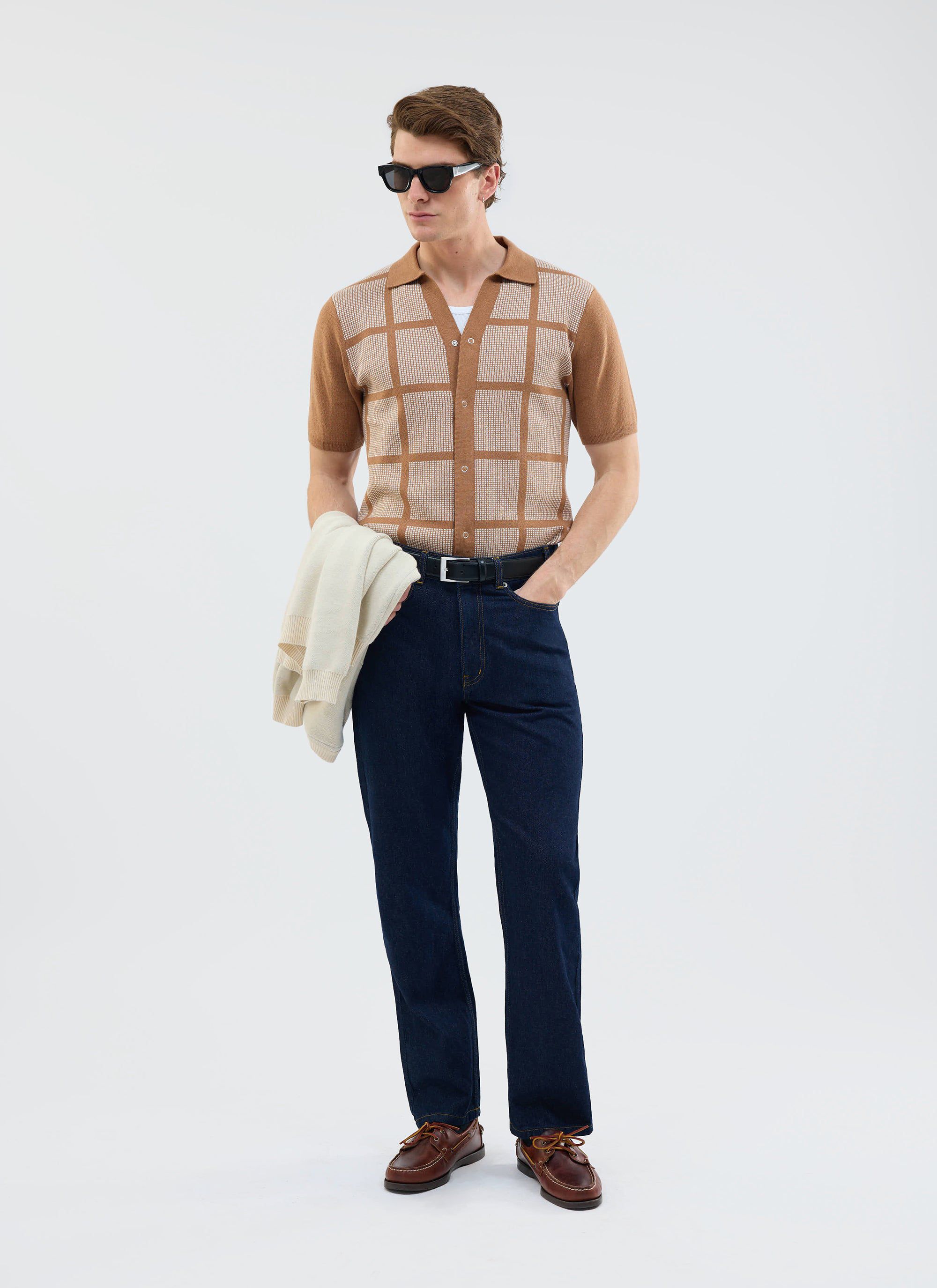 A model wears the Percival Costa Knitted Shirt in tobacco with jeans, boat shoes, and sunglasses against a studio backdrop.