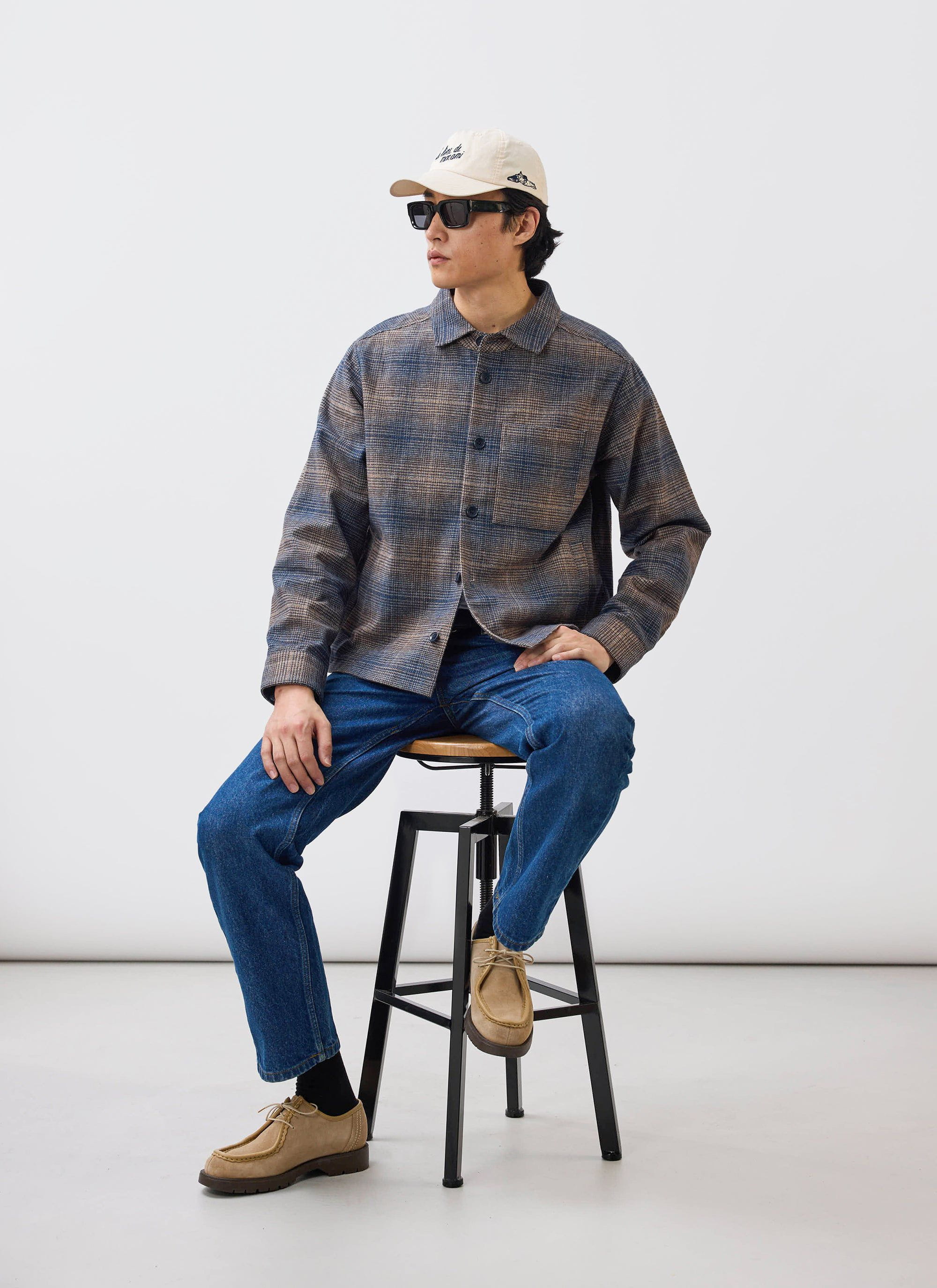 A model wears the Percival Luton Check Overshirt in brown, styled with blue jeans, a cap and sunglasses while sat on a stool.
