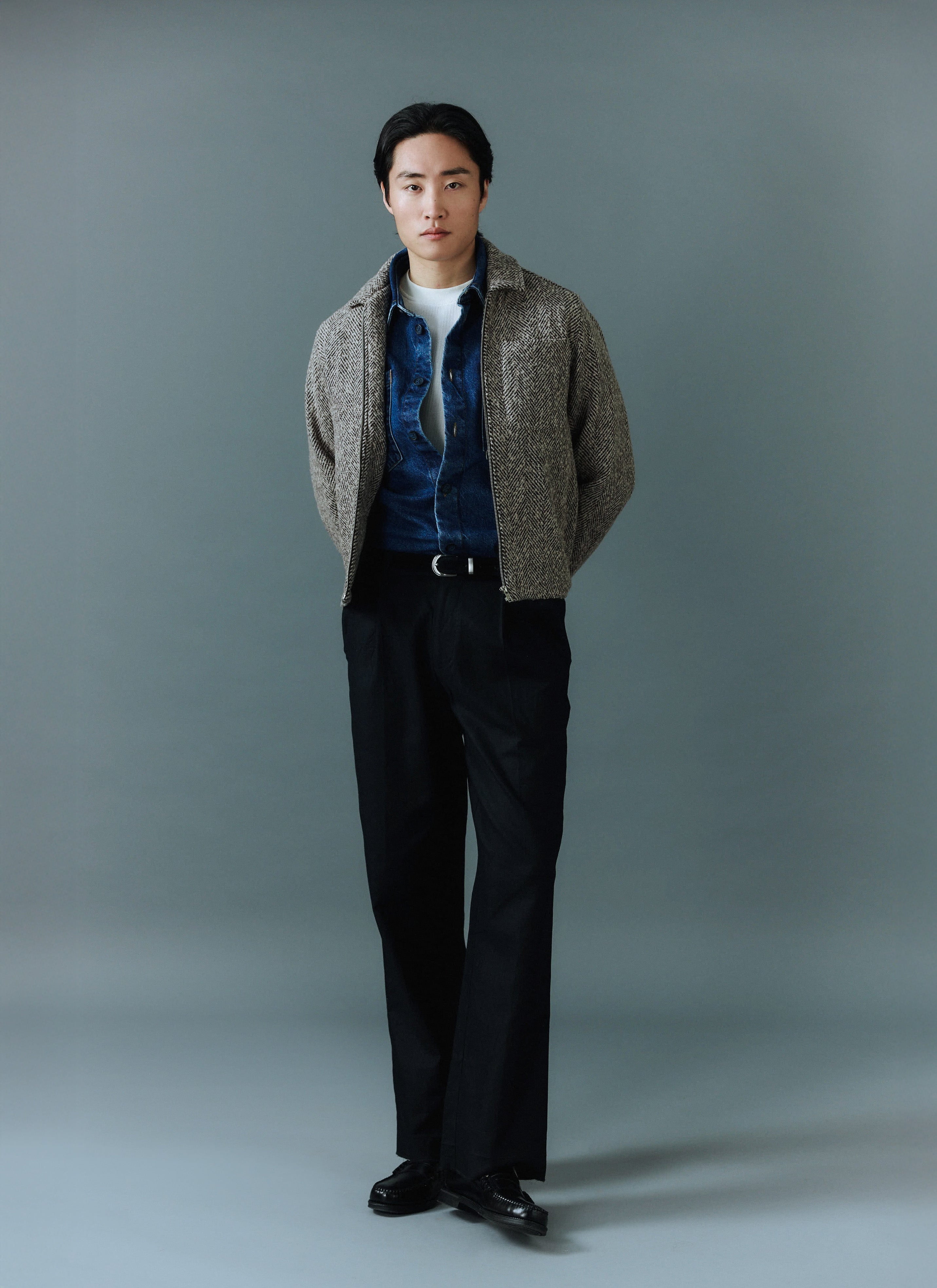A man models the Percival Herringbone Zip Jacket in ecru, layered over a blue denim shirt and paired with black trousers.