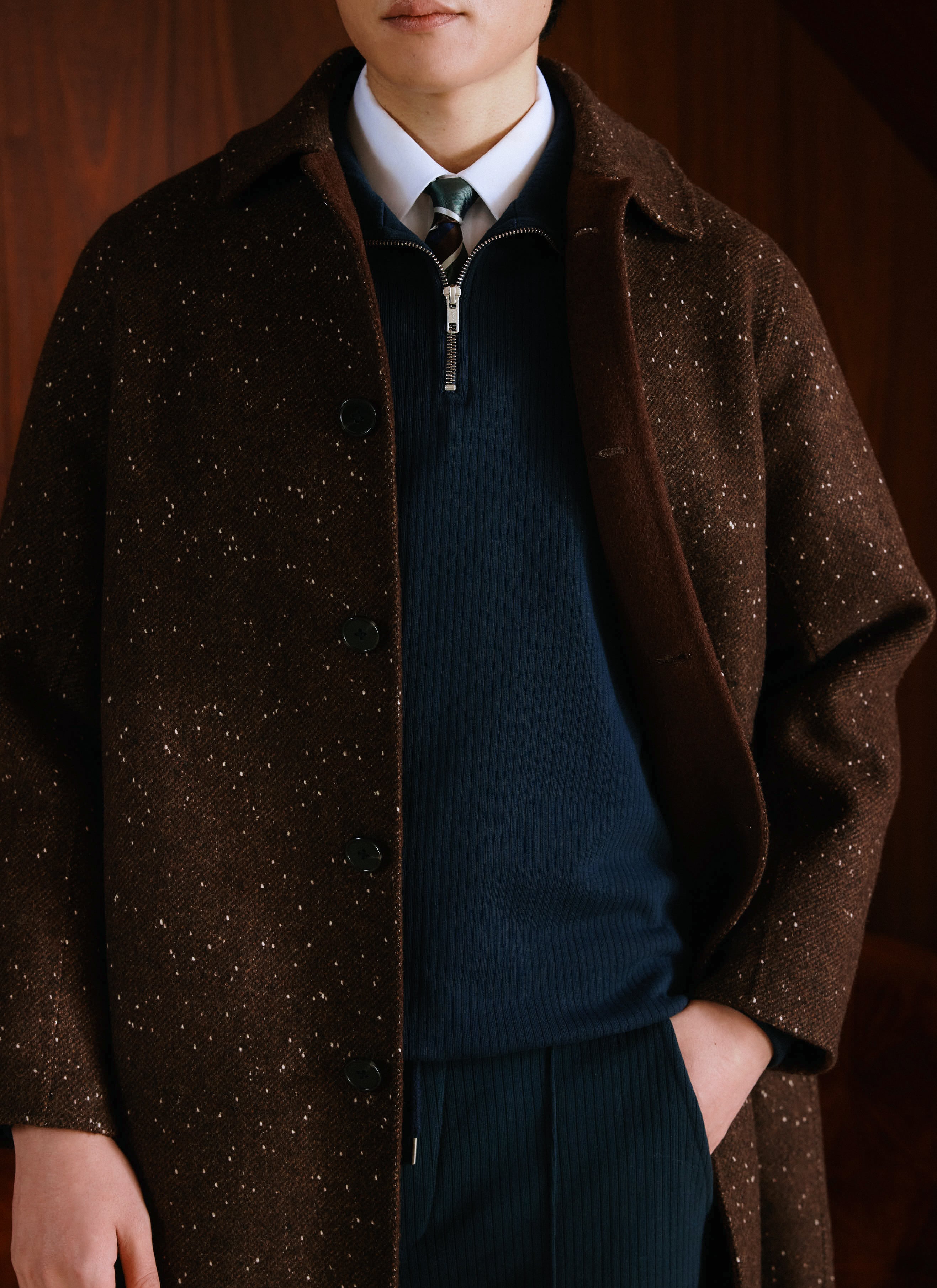 A model wears a Percival brown fleck overcoat over a navy zip jumper, white shirt and tie in a close-up studio shot.