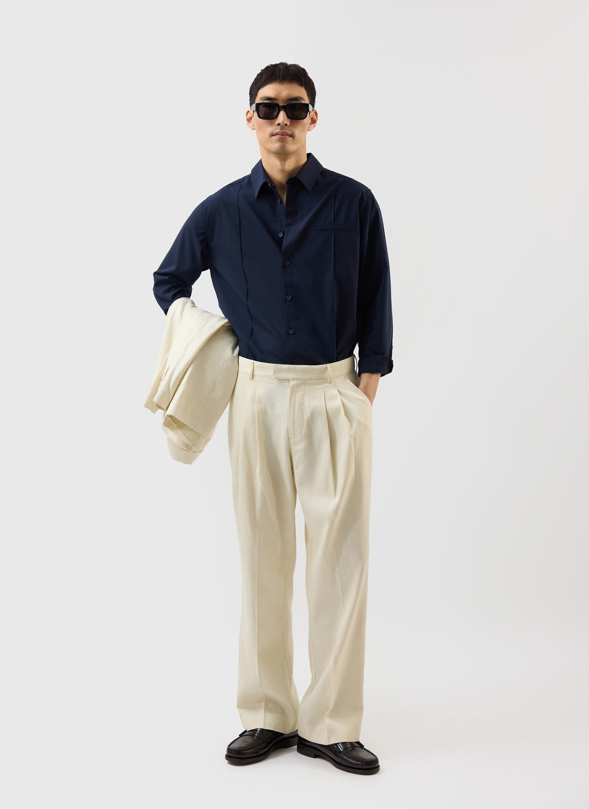 A model wears the Percival x Warren navy pleated shirt with cream trousers and sunglasses, holding a matching cream jacket.