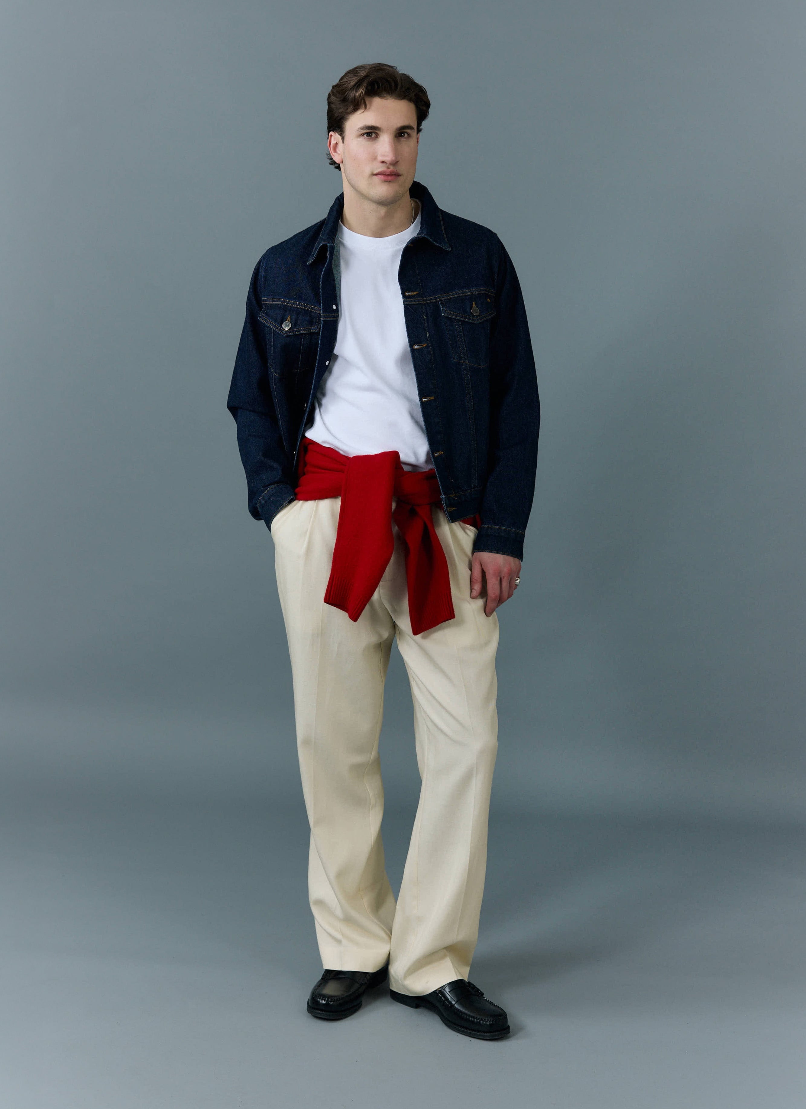 A man models the Percival dark indigo Western Trucker Jacket, styled with a white t-shirt, cream trousers and a red jumper.