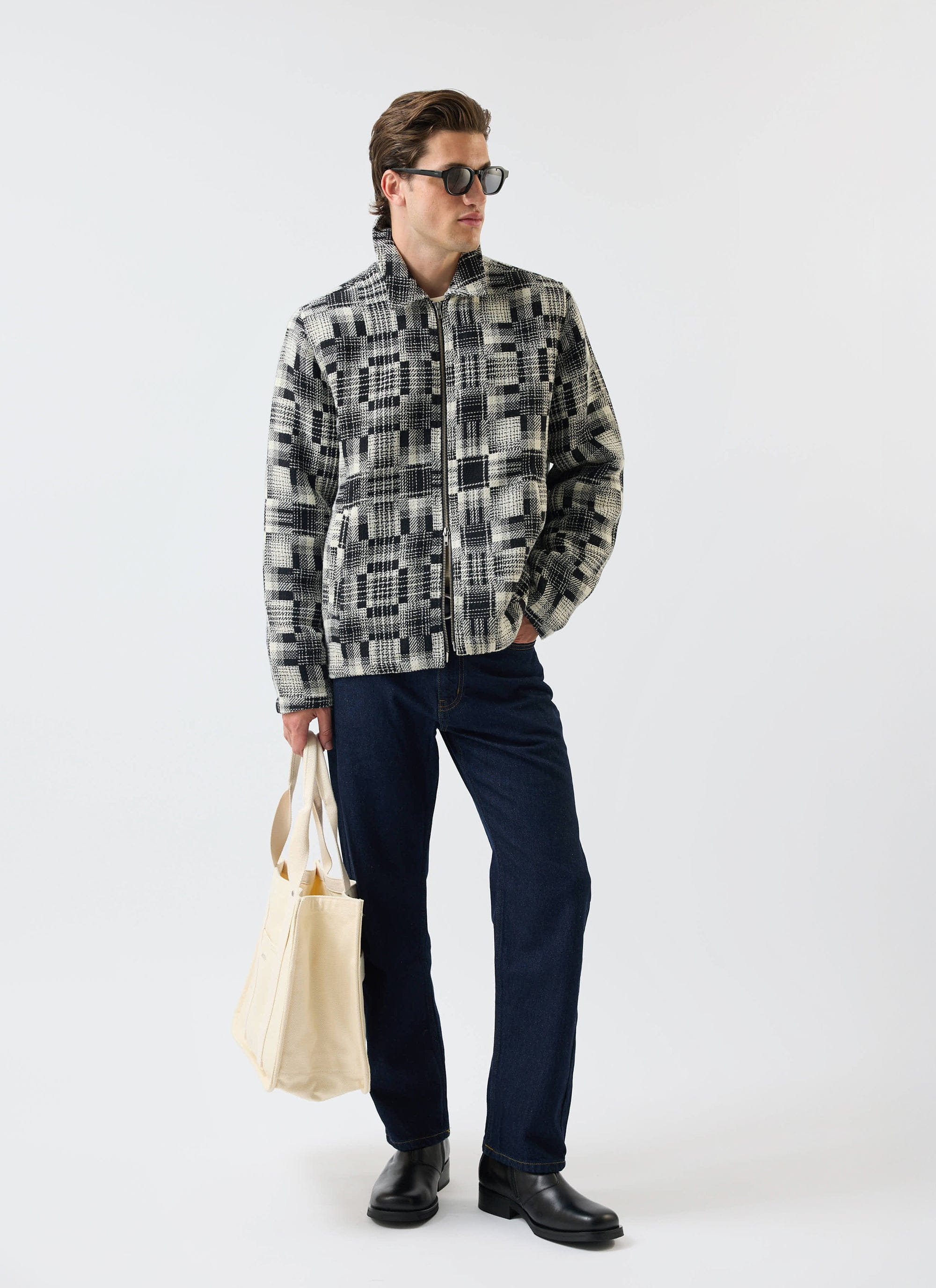 A model wears the Percival Glossary Patchwork Jacket in grey twill, styled with dark denim, sunglasses, and a tote bag.