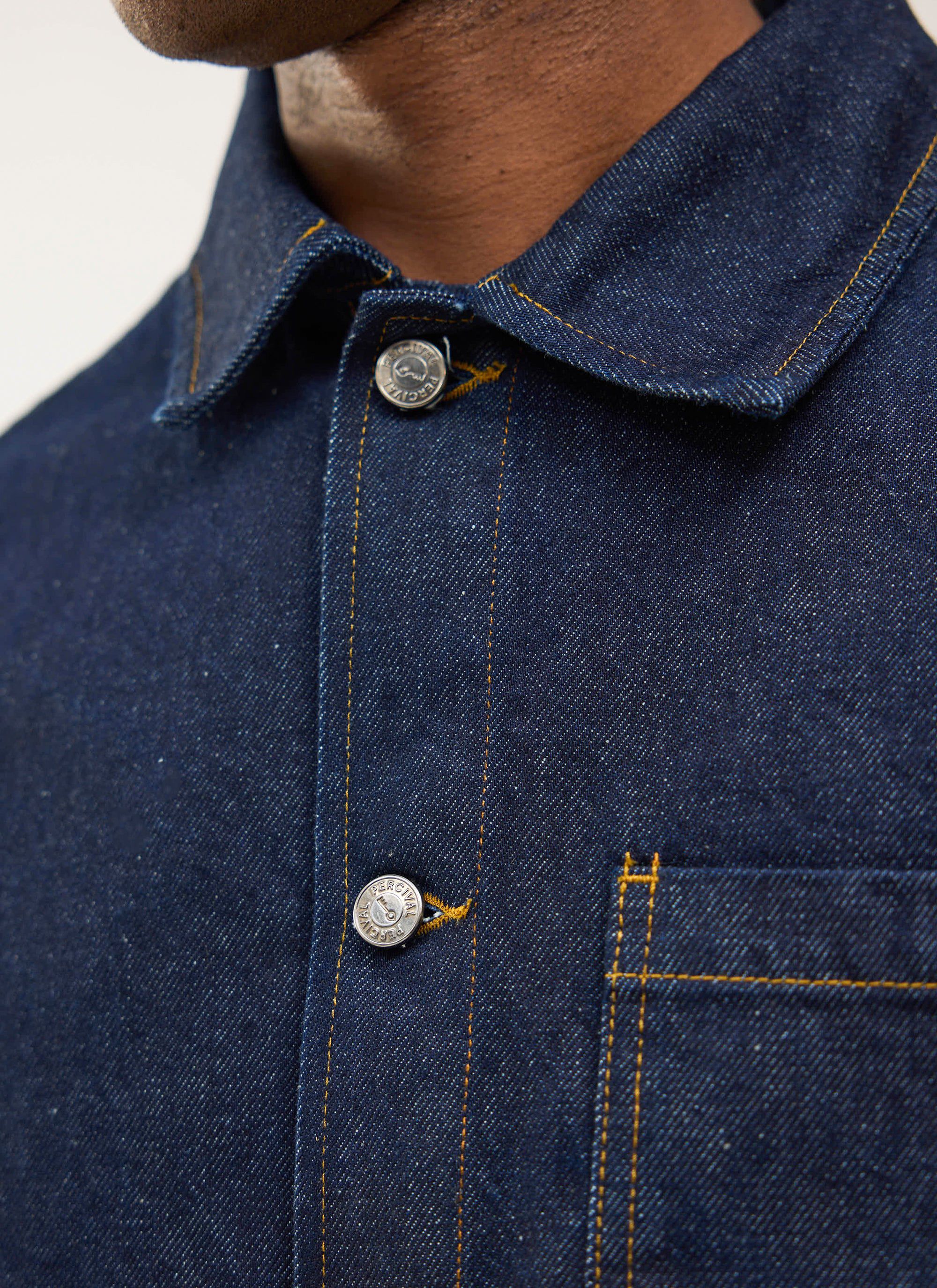 Close-up of the Percival Gove Chore Jacket in dark indigo denim, showing the contrast stitching and branded metal buttons.