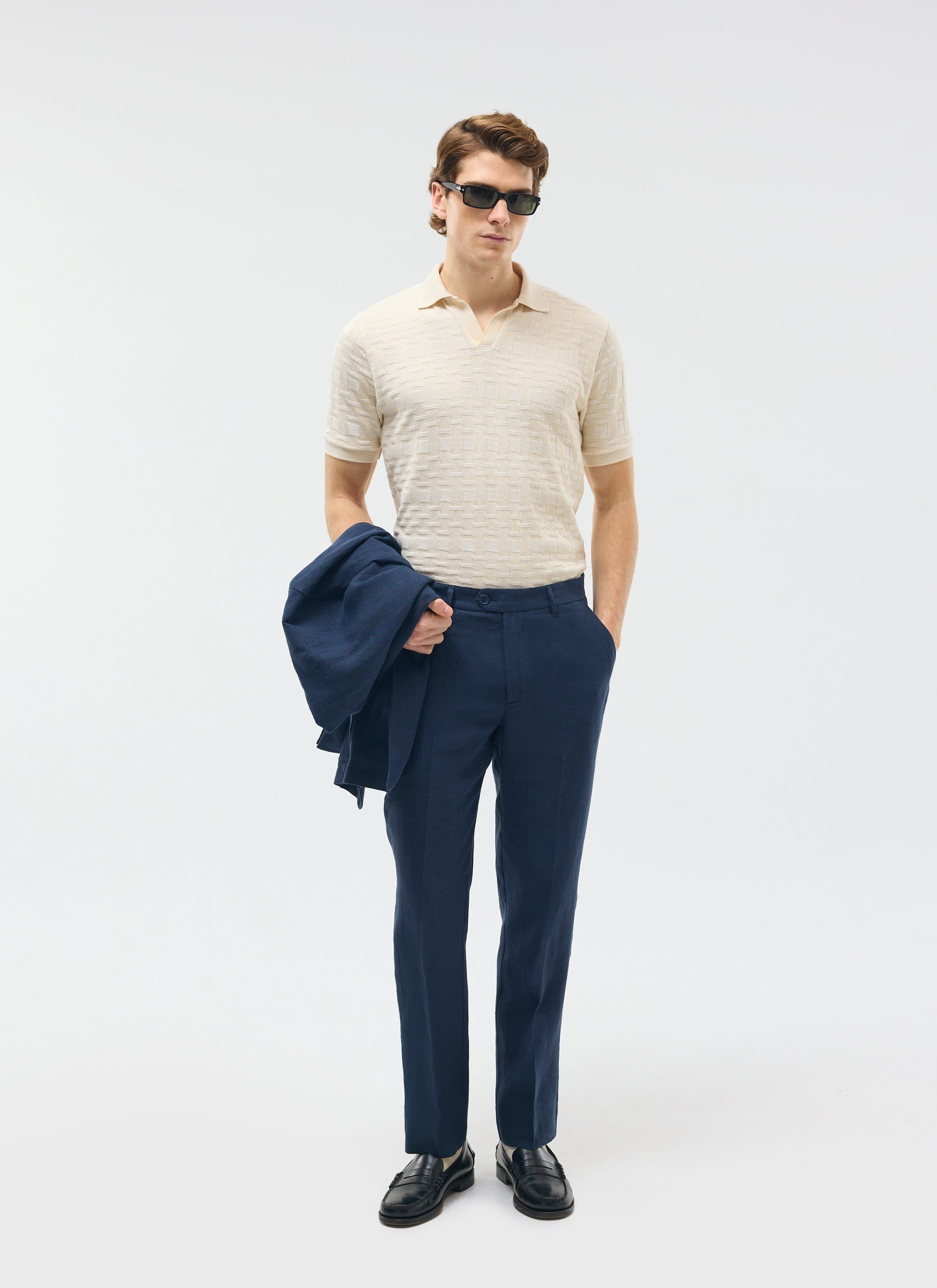 A model wears Percival's navy tailored linen trousers with a cream polo shirt, holding the matching navy jacket.