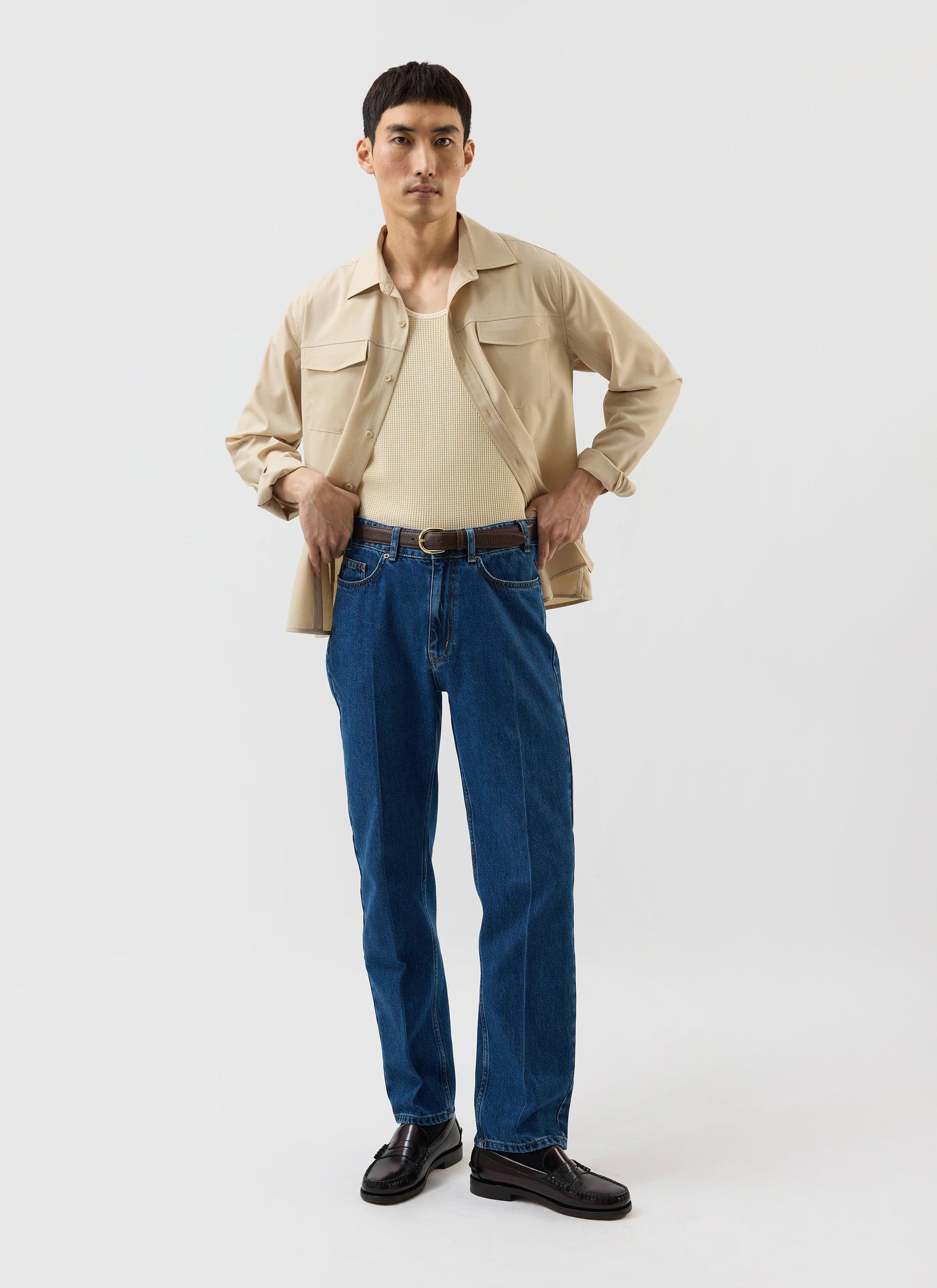 A man models the Percival x Warren Safari Shirt in camel beige, worn open over a vest with blue jeans and loafers.