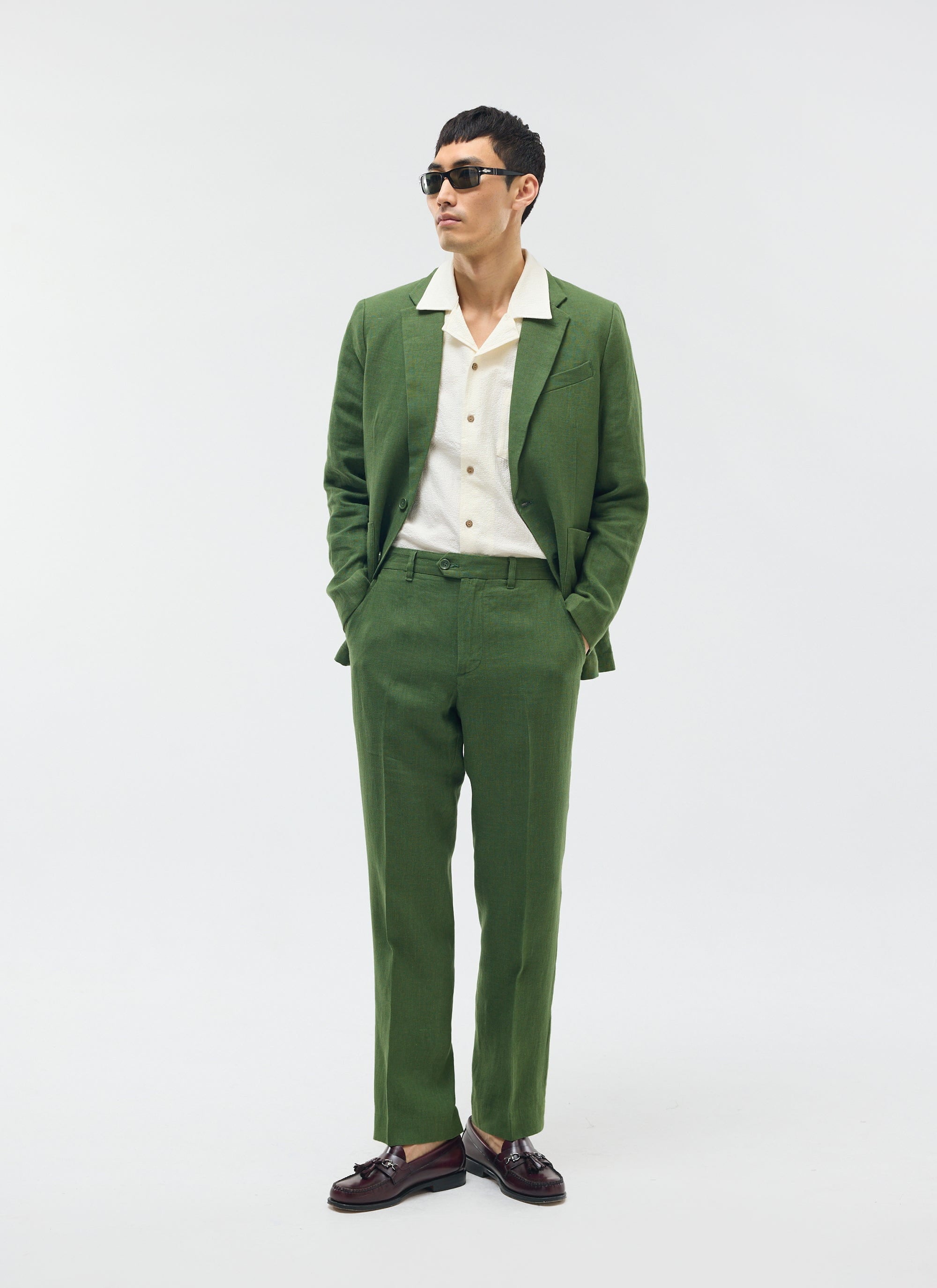 A model wears a Percival forest green tailored linen suit with a cream shirt and sunglasses against a white background.