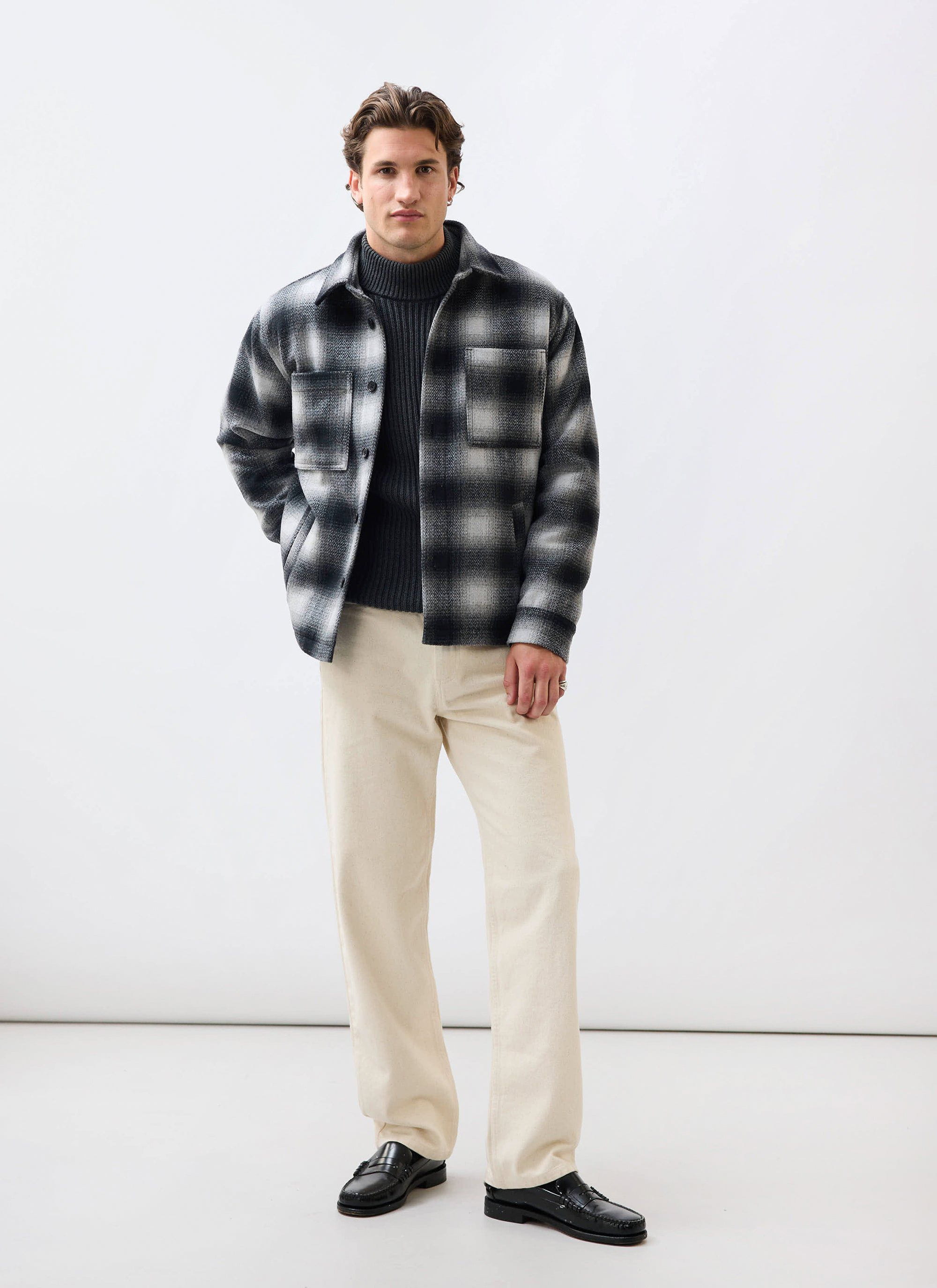 A man models the Percival Check Overshirt in grey, styled with a dark grey turtleneck, cream trousers and black loafers.