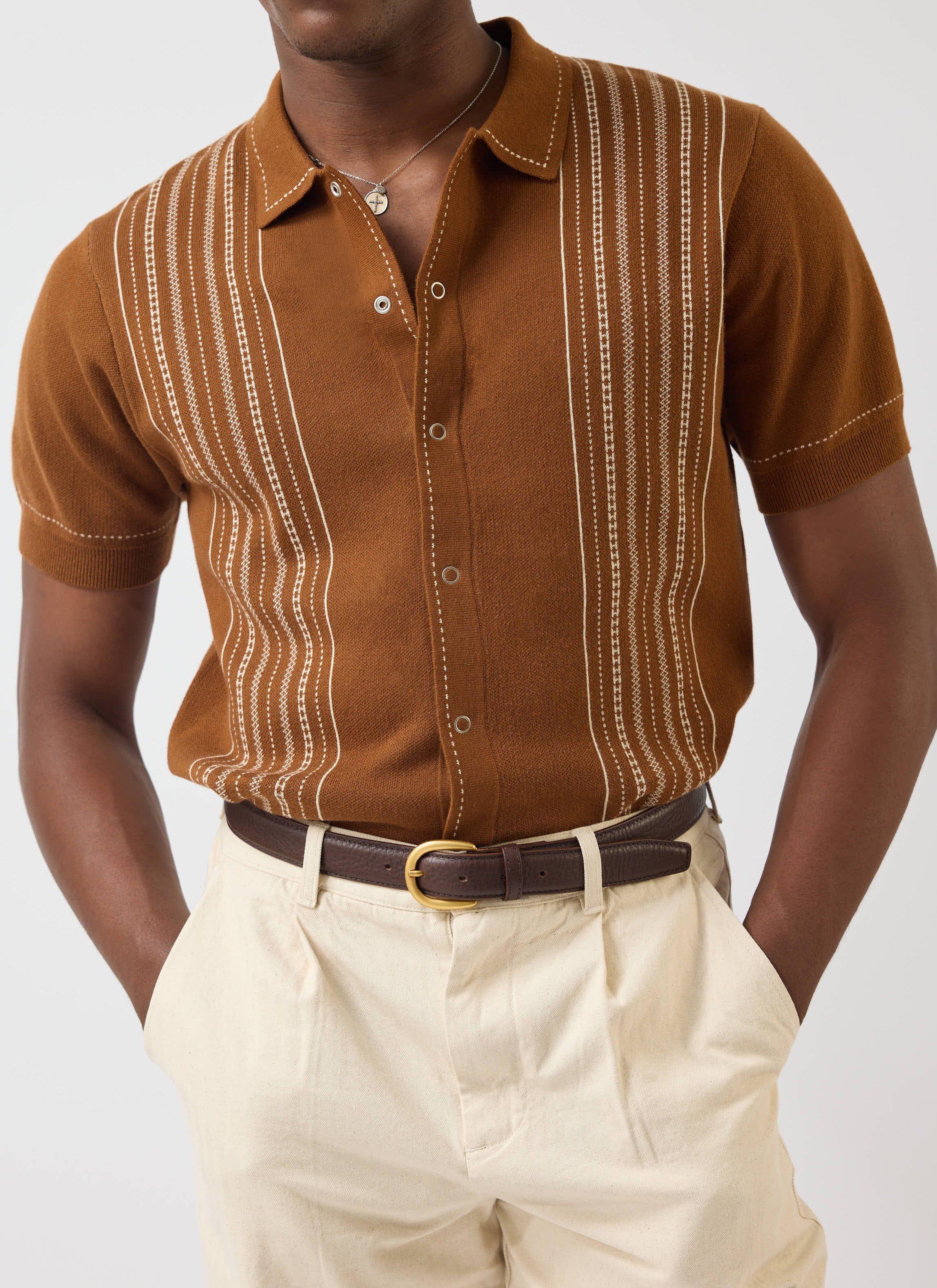 A man models the Percival Accolades Knitted Short Sleeve Shirt in tobacco brown, tucked into cream trousers.
