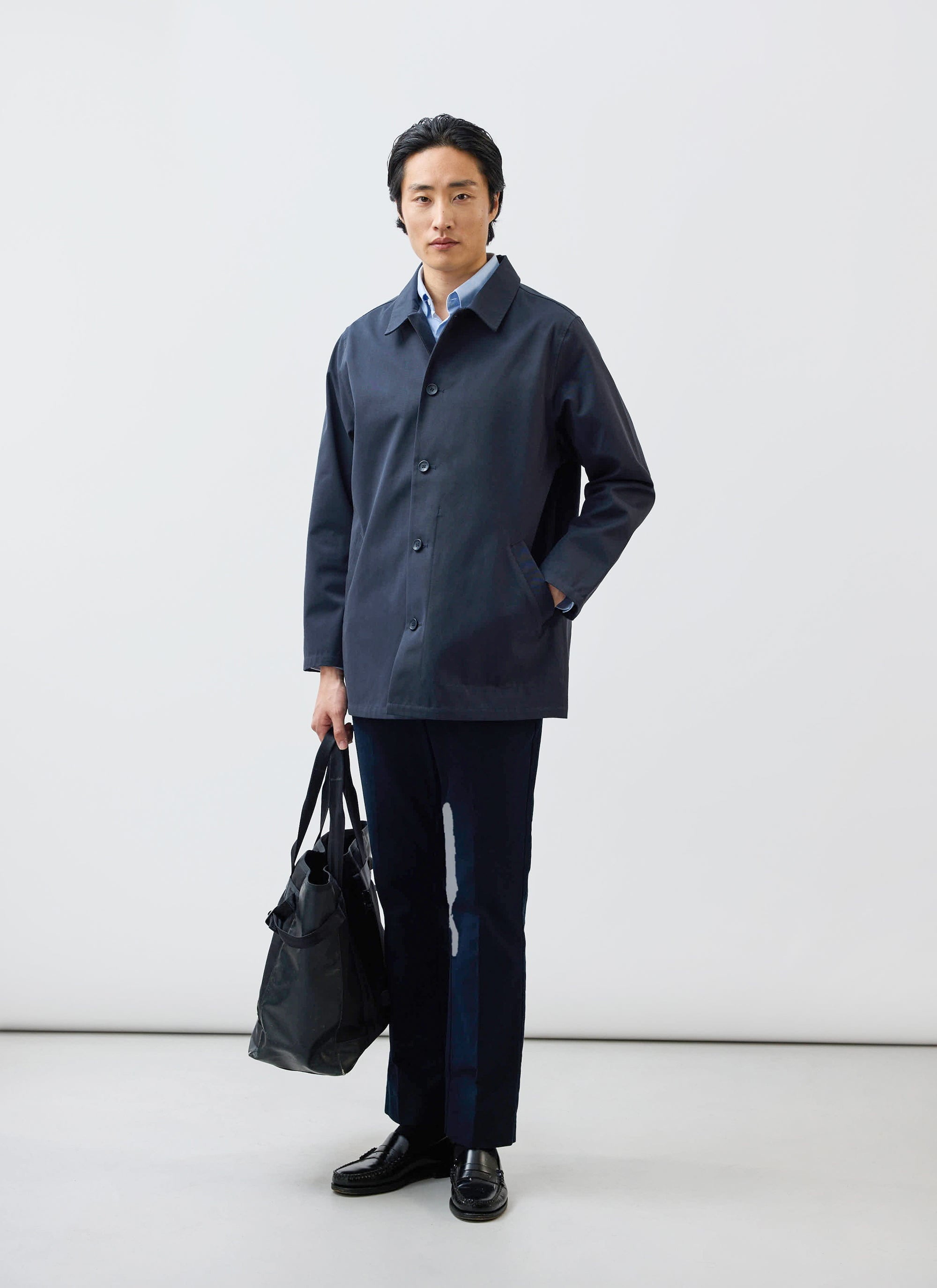 A model wears the Percival navy Sherlock short trench coat with dark trousers and holds a black bag.