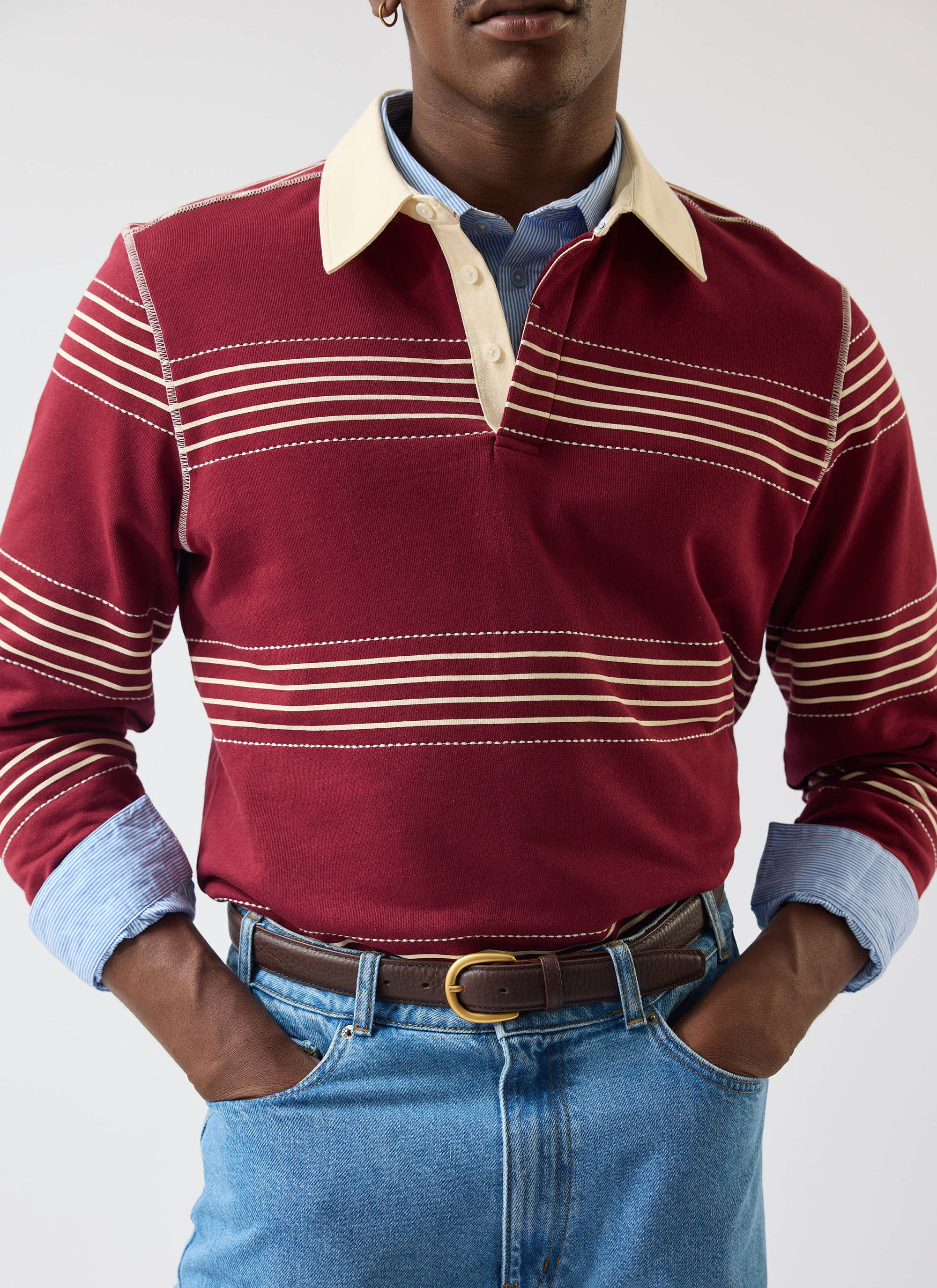 A model wears the Percival Editor Striped Rugby Polo Shirt in burgundy, layered over a blue striped shirt with blue jeans.
