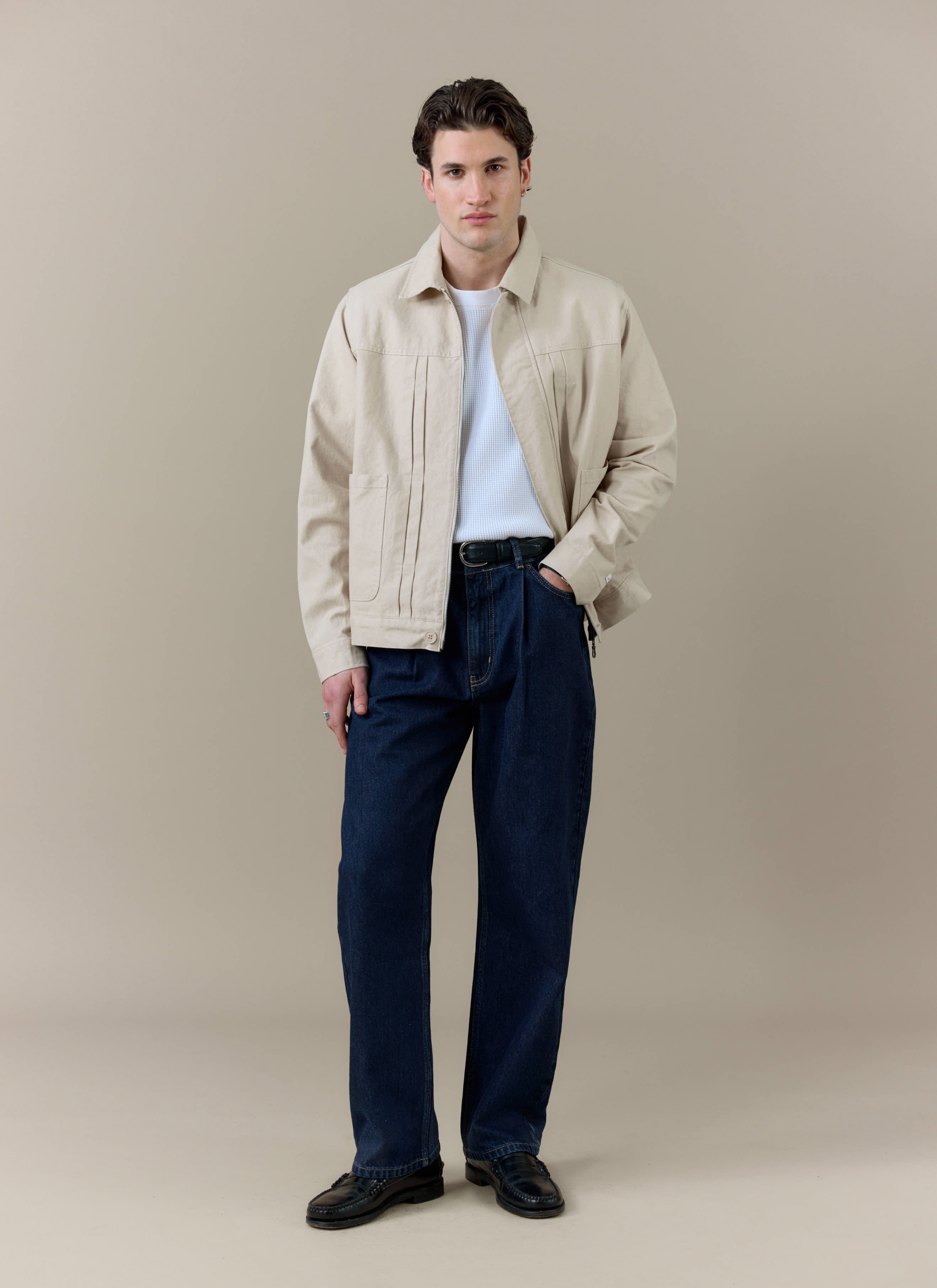 A model wearing the Percival Ernst Jacket in cream cotton, featuring front pleats, styled with dark denim jeans.