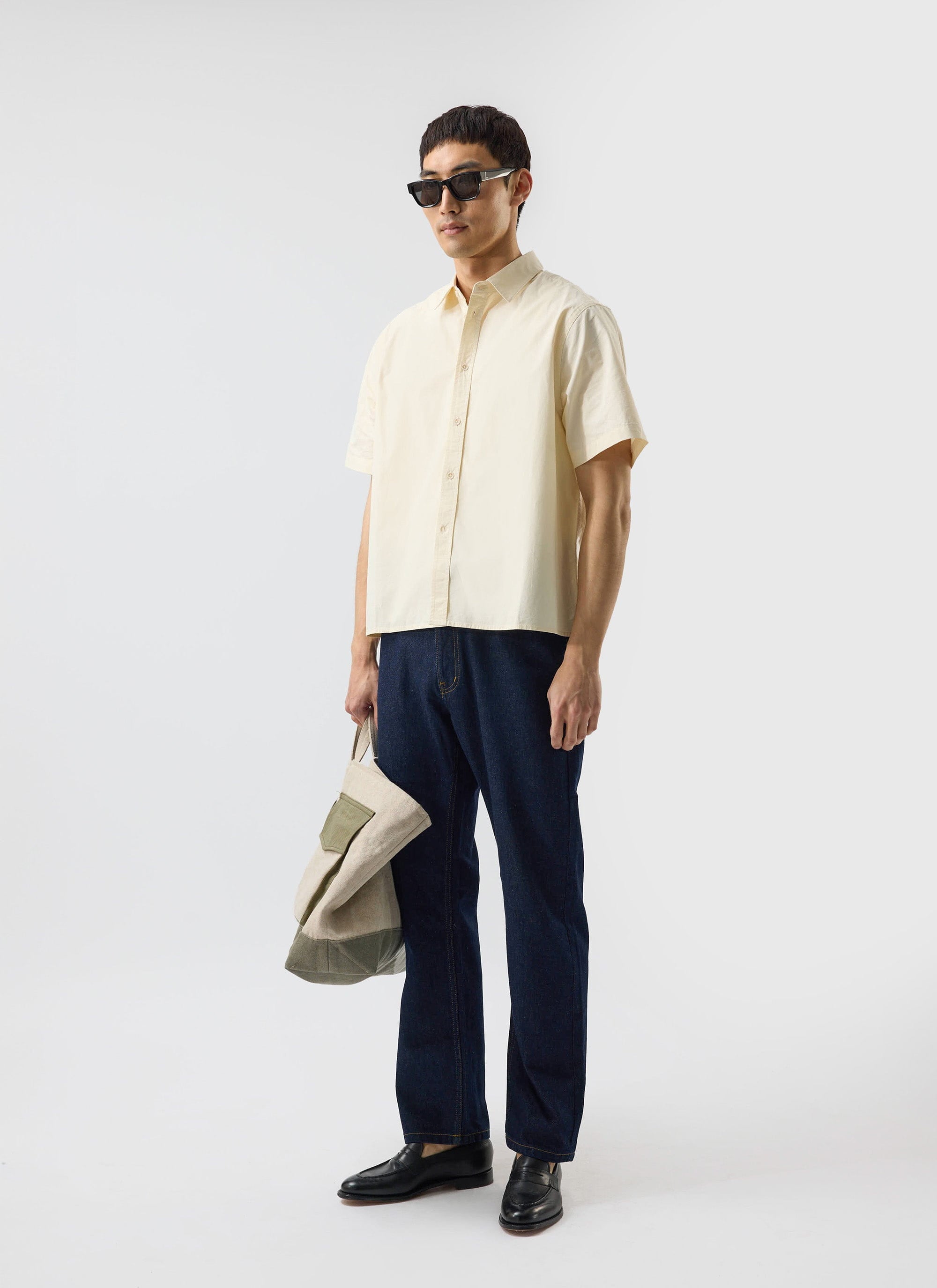 A man models the Percival Boxy Cropped Clerk Shirt in ecru, with dark jeans, black loafers and sunglasses.