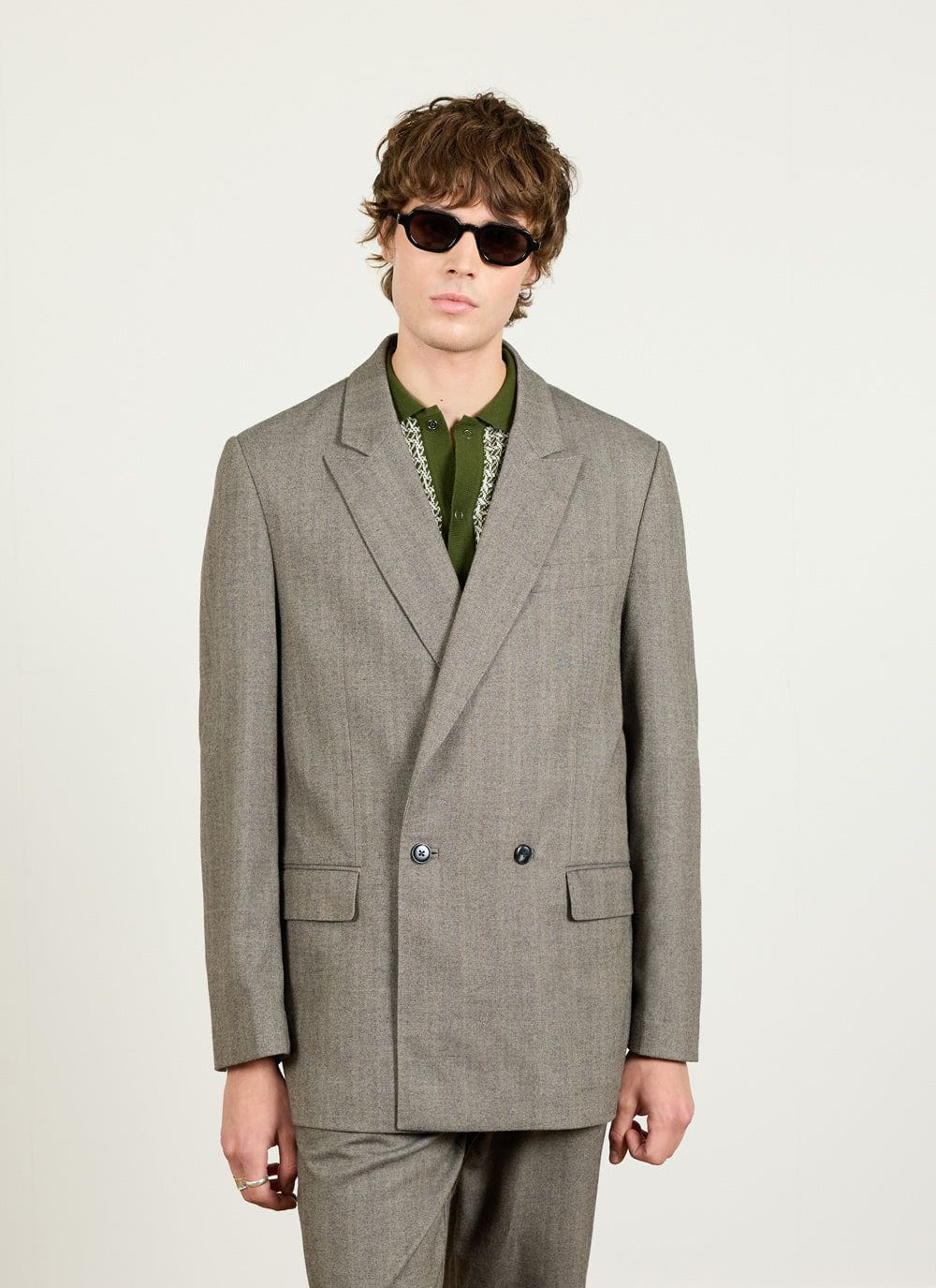 A model wears the Percival grey herringbone double breasted blazer with a green polo shirt and sunglasses.