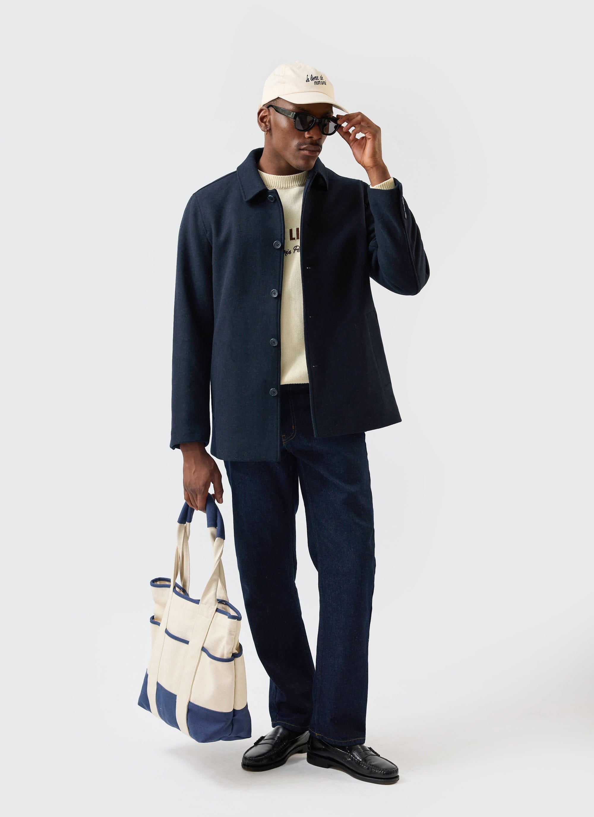 A model wearing the Percival navy wool Peacoat, cream jumper, and dark jeans adjusts his sunglasses while holding a tote bag.