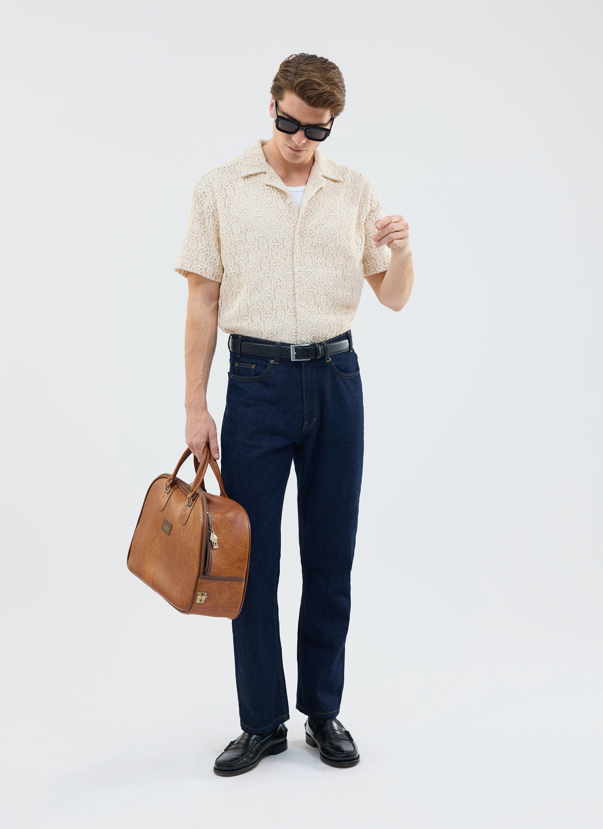 A model wears the Percival Elio Crochet Cuban Shirt in ecru with dark denim, sunglasses, and a leather weekend bag.