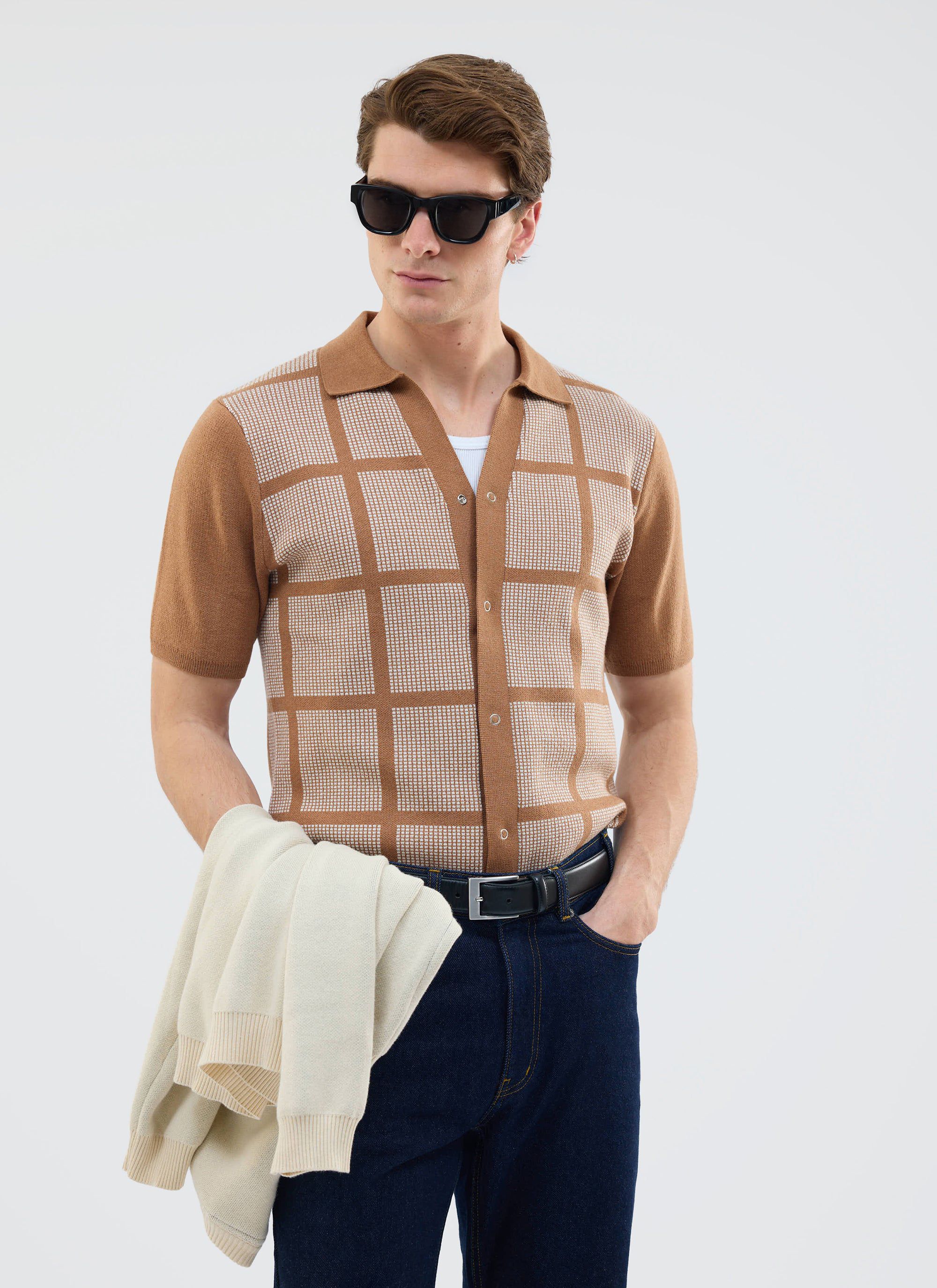 A model wears the Percival Costa Knitted Shirt in tobacco, featuring a square pattern, with dark jeans and sunglasses.